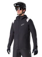 Alpinestars A-Dura Rain Jacket, MTB-regnjakke, sort med lysegrå skulderdetaljer, båret af en rytter med sort MTB-hjelm og solbriller, hvidt logo på brystet, vandtæt konstruktion, koordineret med sorte bukser og handsker til terrænkørsel