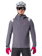 Alpinestars A-Dura Rain Jacket, Mountain Bike Rain Jacket, Dark Titanium Gray, hooded design with pink Alpinestars logo and light gray shoulder panels, worn with white MTB helmet and bright pink gloves, waterproof gear for off-road cycling