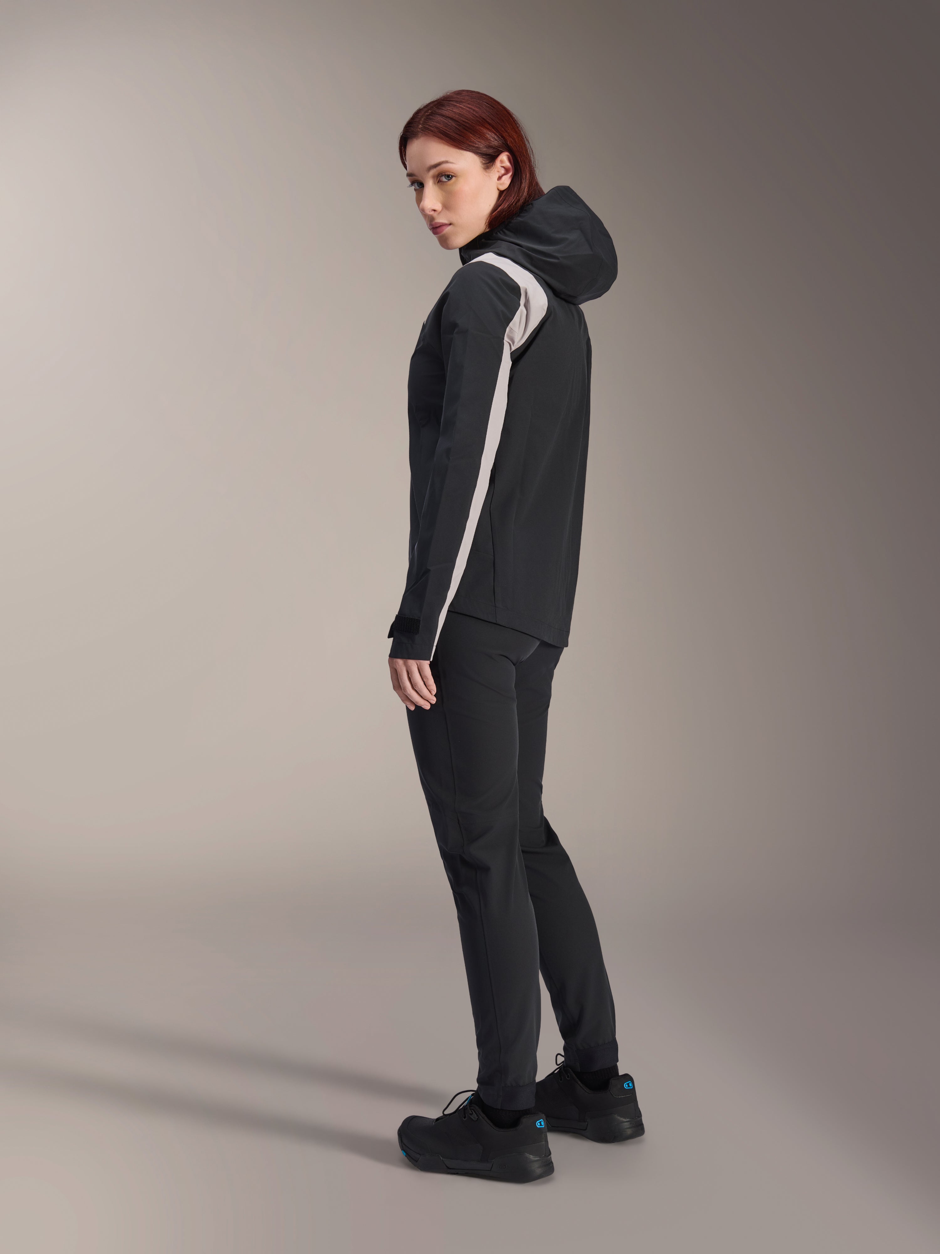 Woman wearing Alpinestars Stella A-Dura Rain Jacket, Black with light gray stripe on the sleeve, hooded waterproof cycling jacket for MTB, paired with black technical pants and black riding shoes, studio profile view