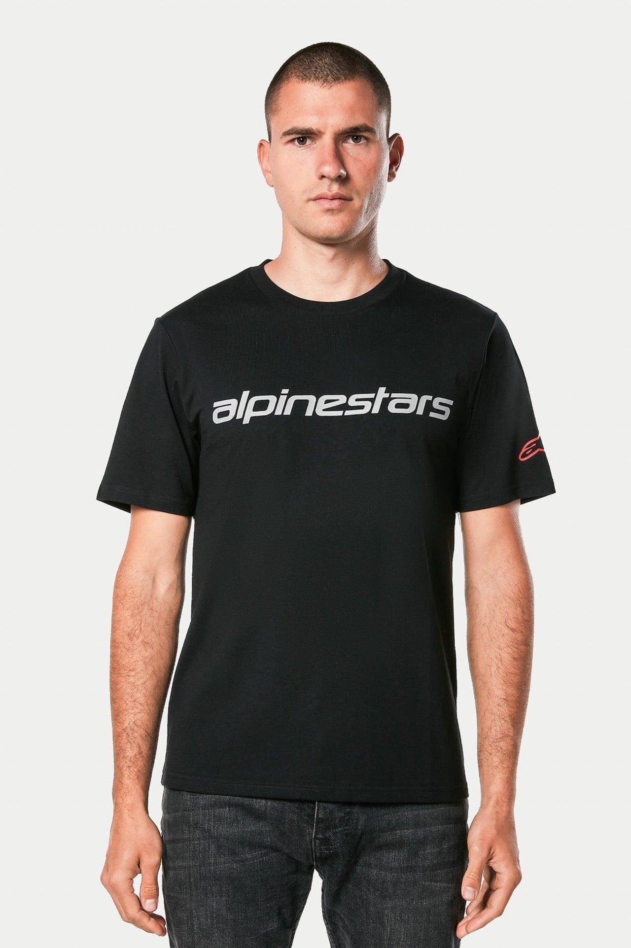 Alpinestars Linear Wordmark 2.0 CSF Tee, sort kortærmet T-shirt, gråt Alpinestars-ordmærke på brystet, rødt Ageless-logo på ærmet, båret af model med mørke jeans, afslappet motorsport-streetwear