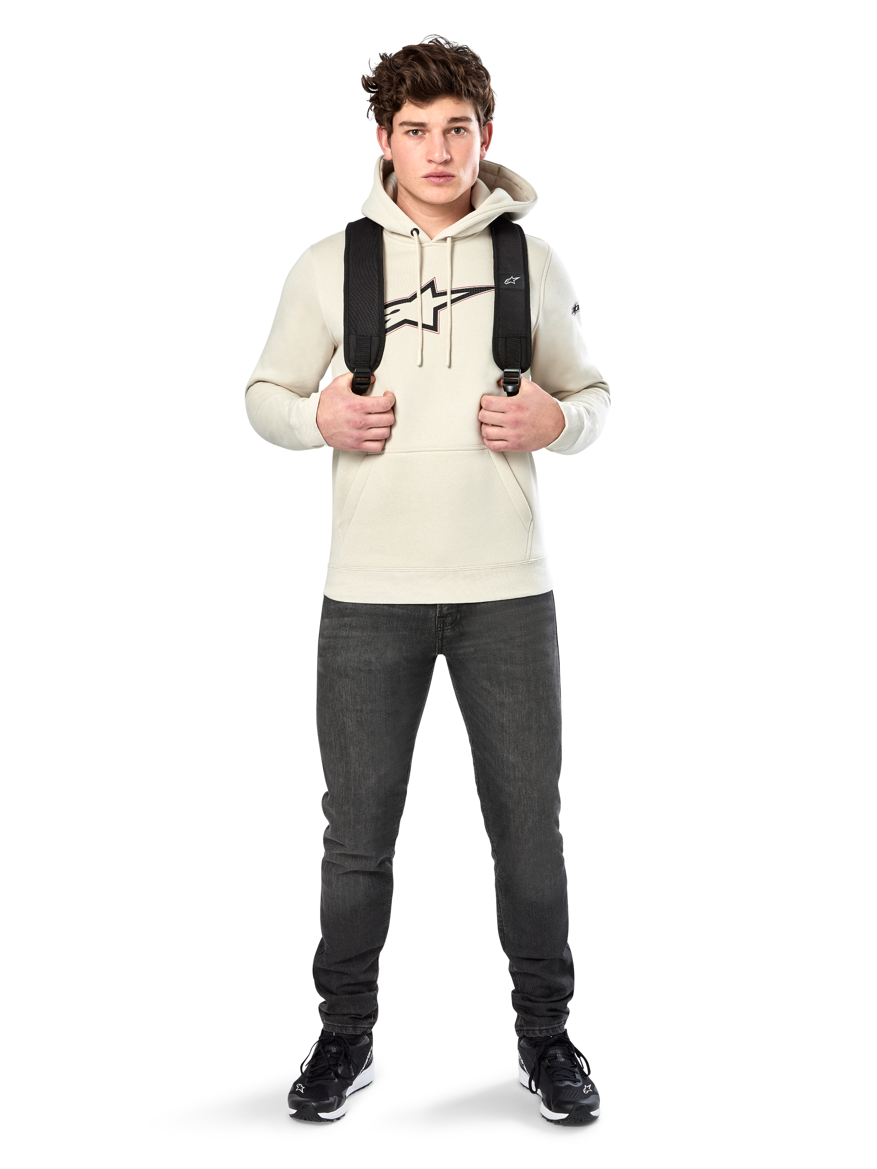 Alpinestars Insignia Hoodie, casual streetwear, Off White fleece pullover, carbon fiber look logo with red outline, drawstring hood, kangaroo pocket, worn by model with black backpack and dark grey jeans
