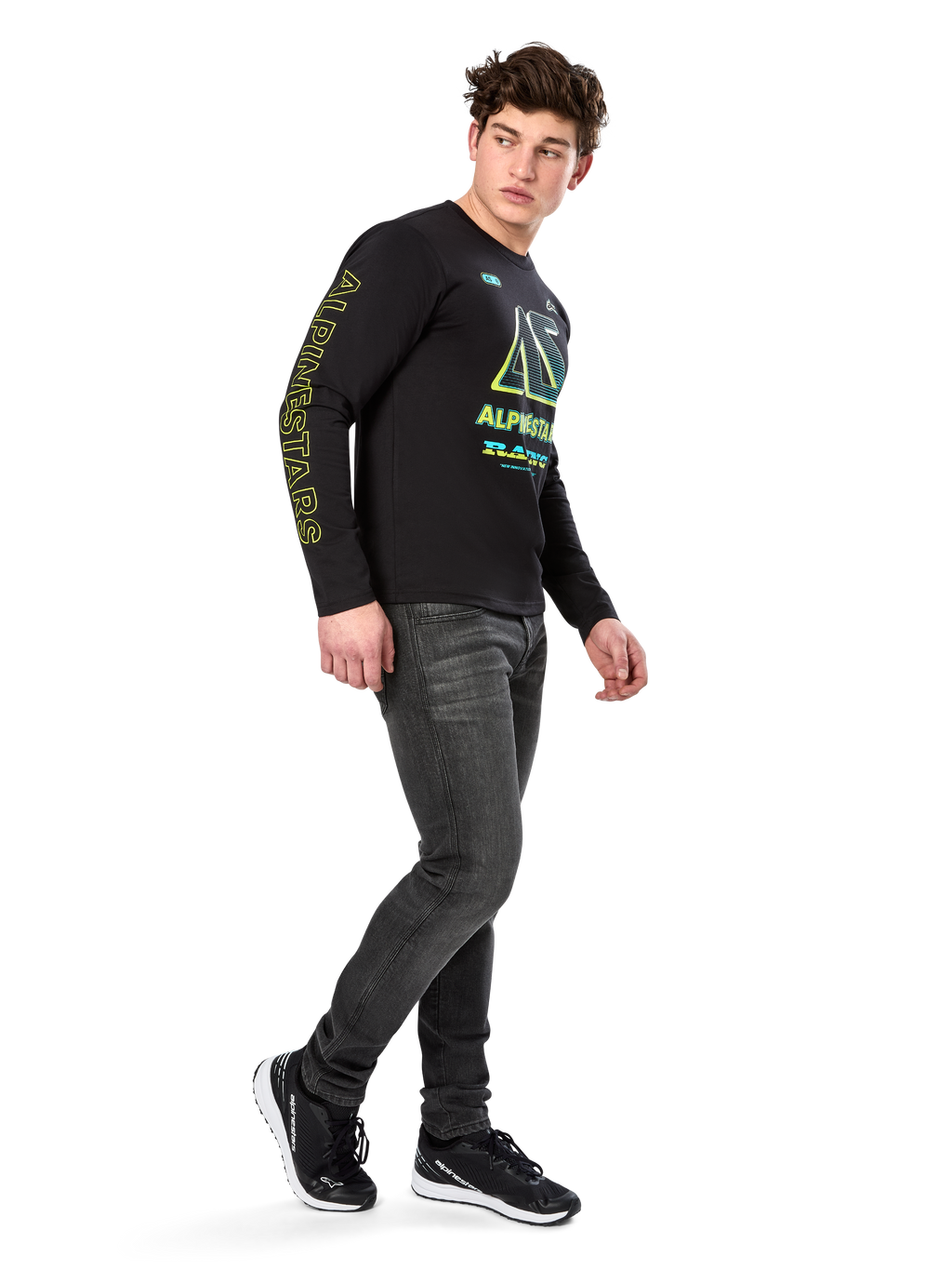 Mand iført Alpinestars Ayess CSF langærmet T-shirt, sort, med gul skrift på ærmet og levende limegrøn og cyan grafik på brystet, kombineret med mørkegrå jeans og sorte sneakers, afslappet motorsport-inspireret streetwear.