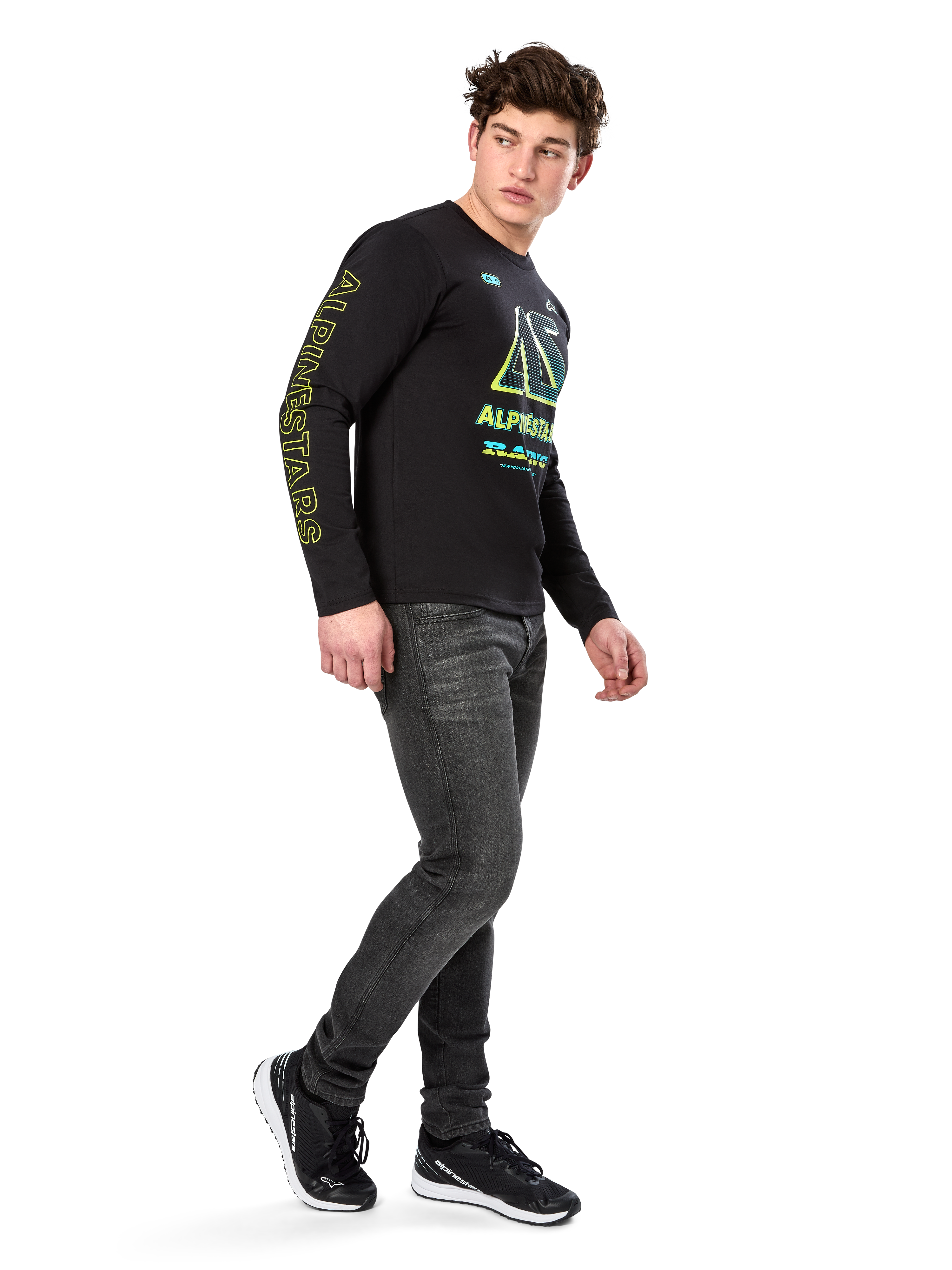 Mand iført Alpinestars Ayess CSF langærmet T-shirt, sort, med gul skrift på ærmet og levende limegrøn og cyan grafik på brystet, kombineret med mørkegrå jeans og sorte sneakers, afslappet motorsport-inspireret streetwear.