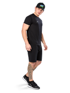 Man with tattoos wearing Alpinestars Point Performance Tee, Short sleeve black performance t-shirt, small white logo, paired with black shorts, a black cap with colorful graphic, and black and white sneakers, sporty casual lifestyle