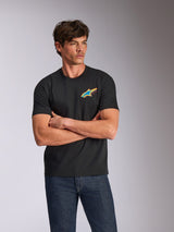 Alpinestars Angler CSF Tee, kortærmet T-shirt, sort, med et livligt flerfarvet Astars-logo på brystet, båret af en model med krydsede arme og blå jeans, afslappet motorsport-inspireret streetwear