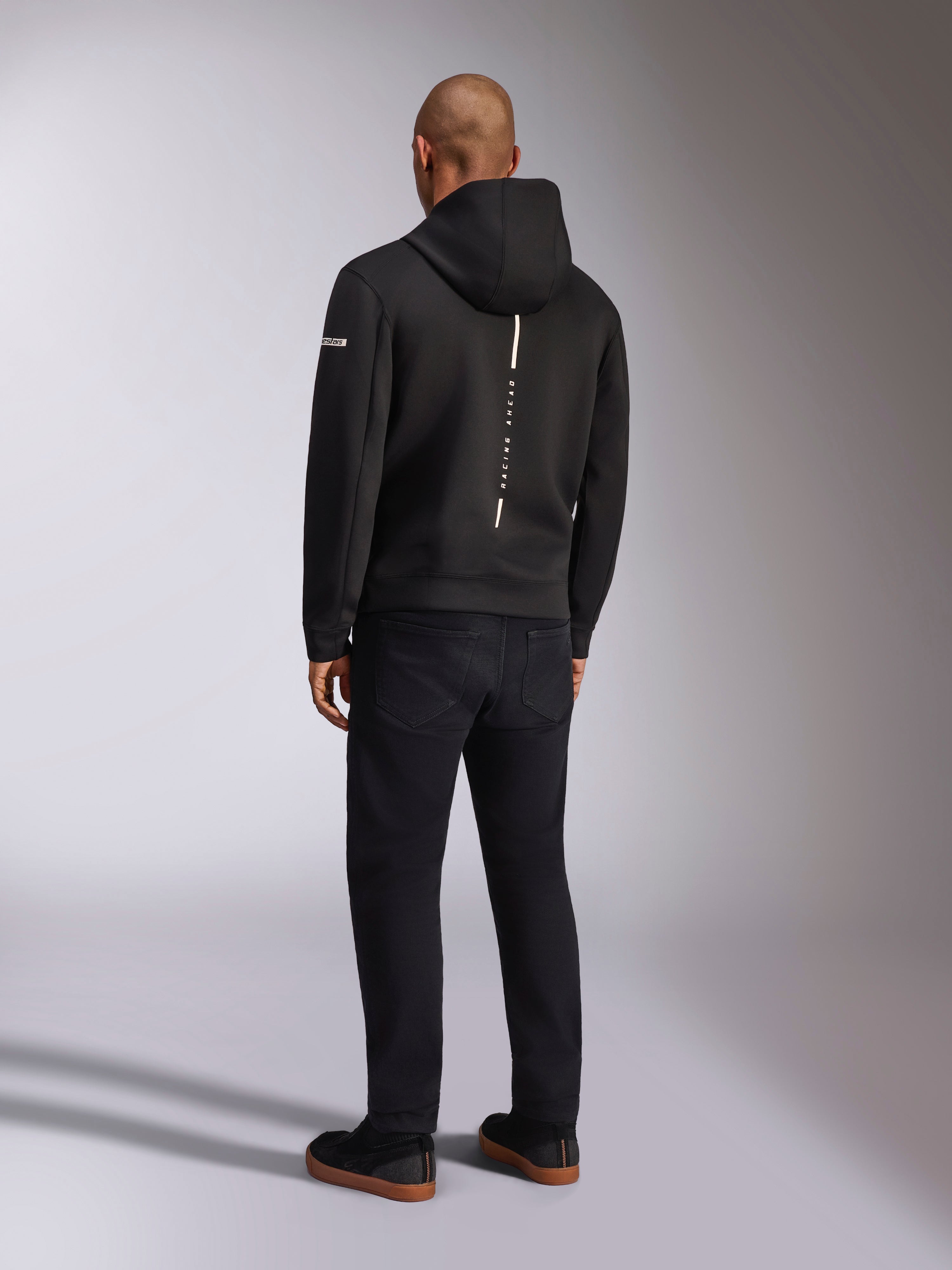 Alpinestars Insight Hættetrøje, sort sportshættetrøje, set bagfra med en lodret hvid stribe med 'RACING AHEAD' branding, logo på ærmet, kombineret med sorte bukser og sneakers, afslappet motorsport-inspireret design