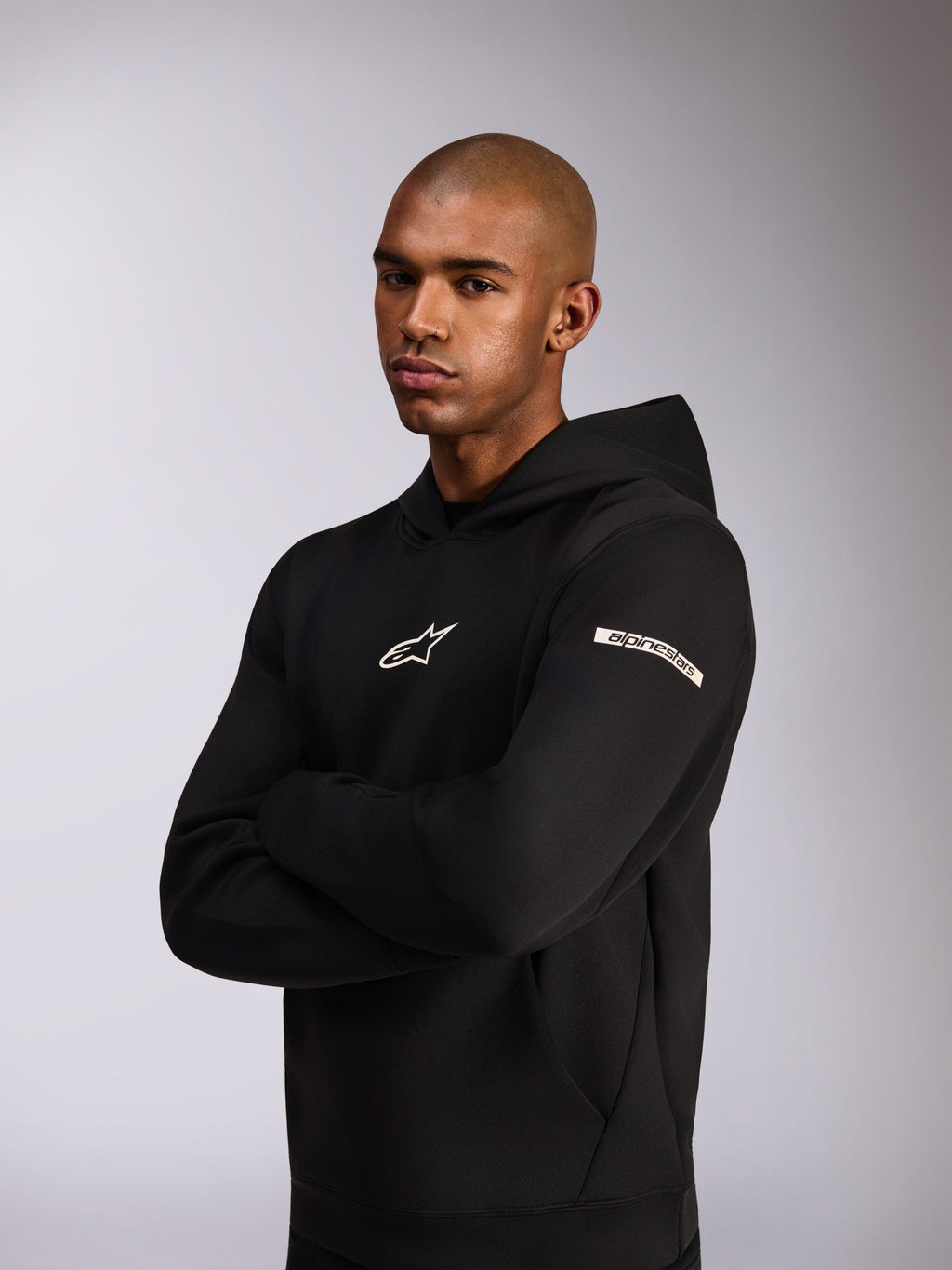 Alpinestars Insight Hoodie, afslappet sportstøj-hættetrøje, sort, hvidt Astars-logo på brystet og tekstlogo på overarmen, model iført hættetrøje med armene over kors, førsteklasses dobbeltsidet fleece-konstruktion