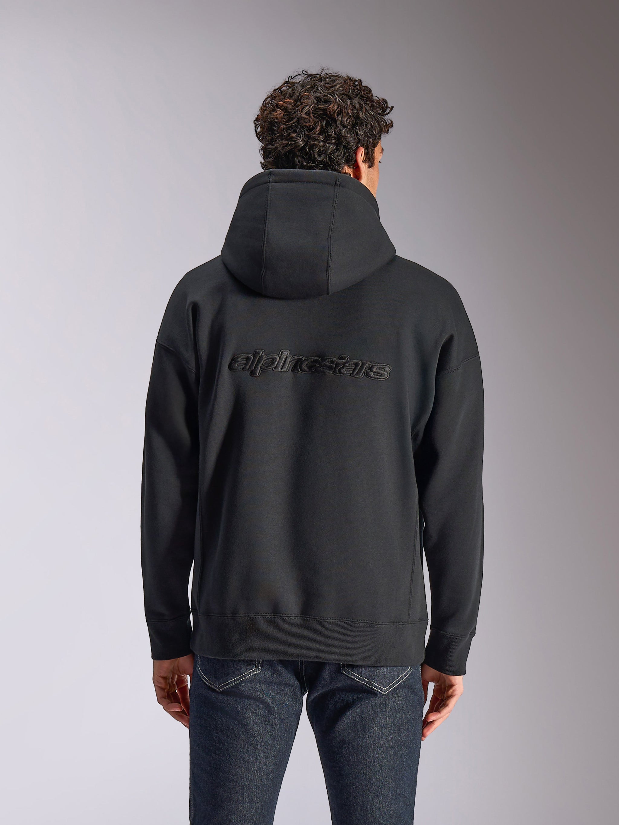 Alpinestars Emergent Oversized Hoodie, bagsidevisning af en sort pullover-hættetrøje på en model, med broderet Alpinestars-ordlogo på ryggen, oversized pasform, afslappet streetwear kombineret med mørke jeans