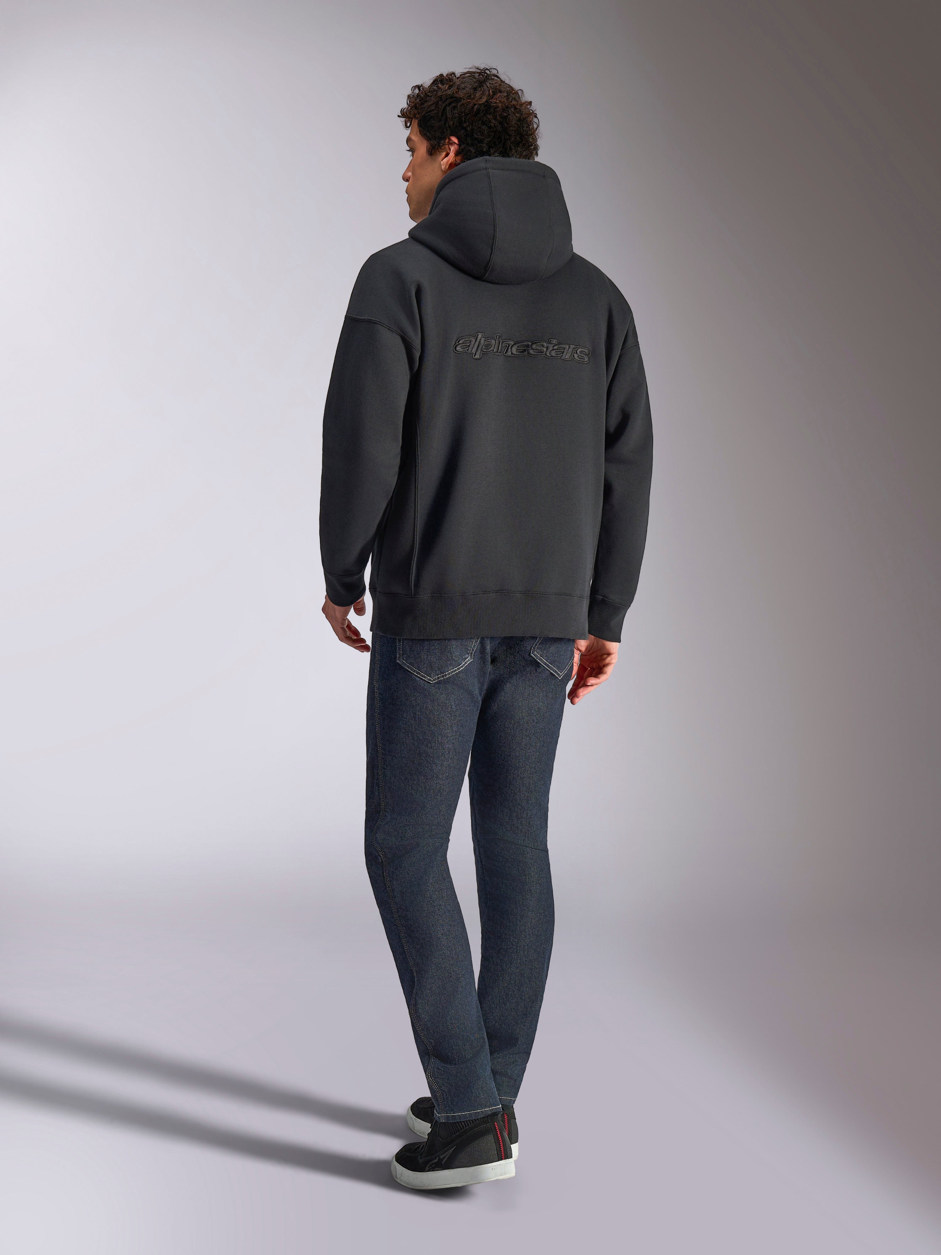 Alpinestars Emergent Oversized Hoodie, Black, back view showing 3D embroidered Alpinestars word logo across the shoulders, oversized fit, styled with dark blue denim jeans and black sneakers, casual motorsport-inspired streetwear.