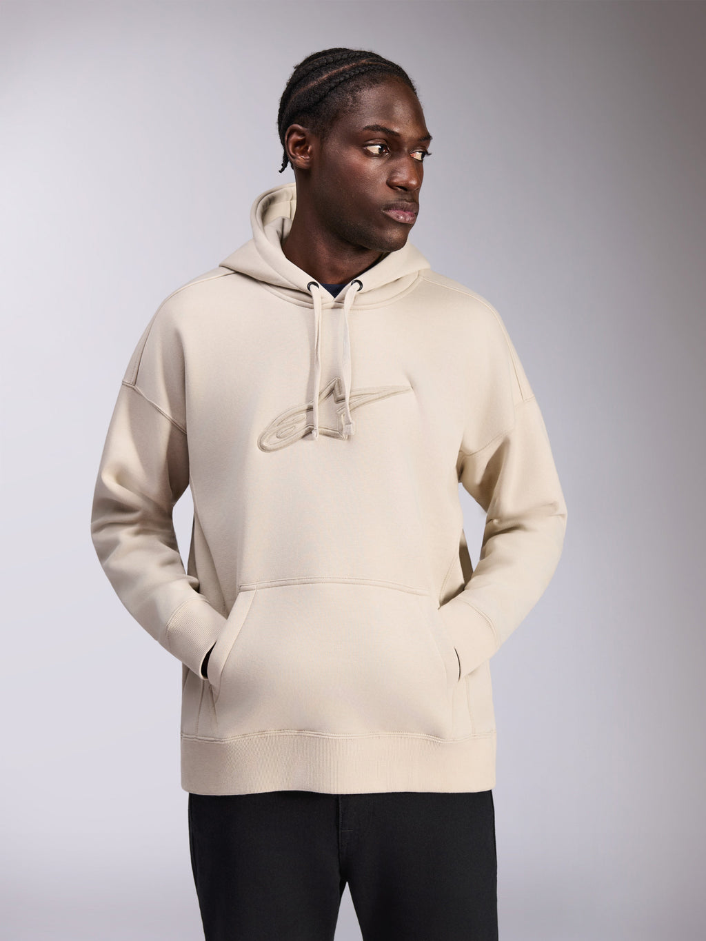 Alpinestars Emergent Oversized hættetrøje, sandfarvet, oversized pullover med tonalt 3D-broderet Astars-logo på brystet, kængurulomme, hætte med snoretræk, afslappet streetwear båret af model med sorte bukser.