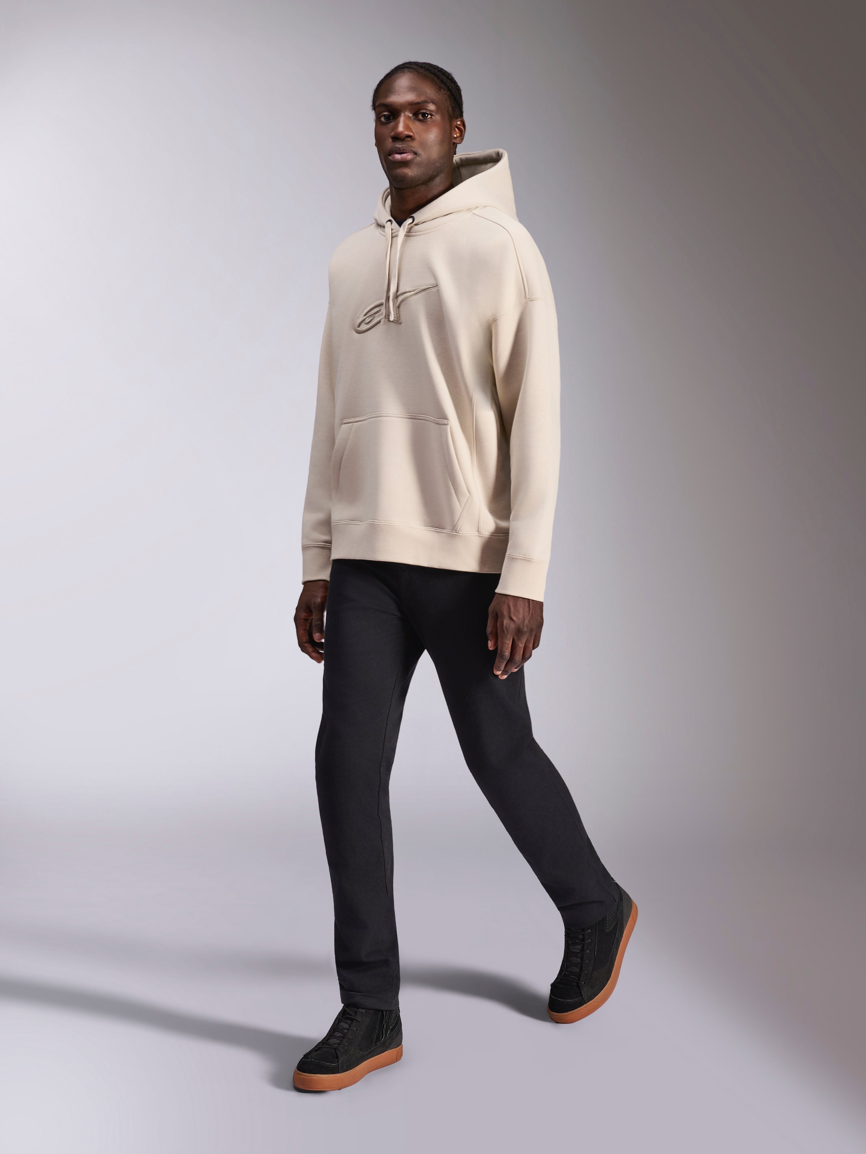 Alpinestars Emergent Oversized hættetrøje, afslappet streetwear, sand off-white, tonalt 3D-broderet logo, båret af model med sorte bukser og sorte sneakers, afslappet oversized pasform med kængurulomme og hætte med snøre