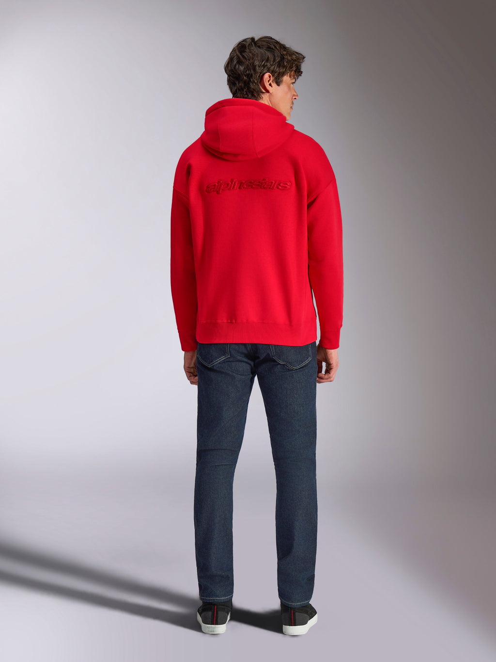 Alpinestars Emergent Oversized Hoodie, rød, set bagfra på mandlig model, med tone-i-tone 3D-broderet Alpinestars ordlogo, båret med mørkeblå jeans, afslappet motorsport-inspireret streetwear