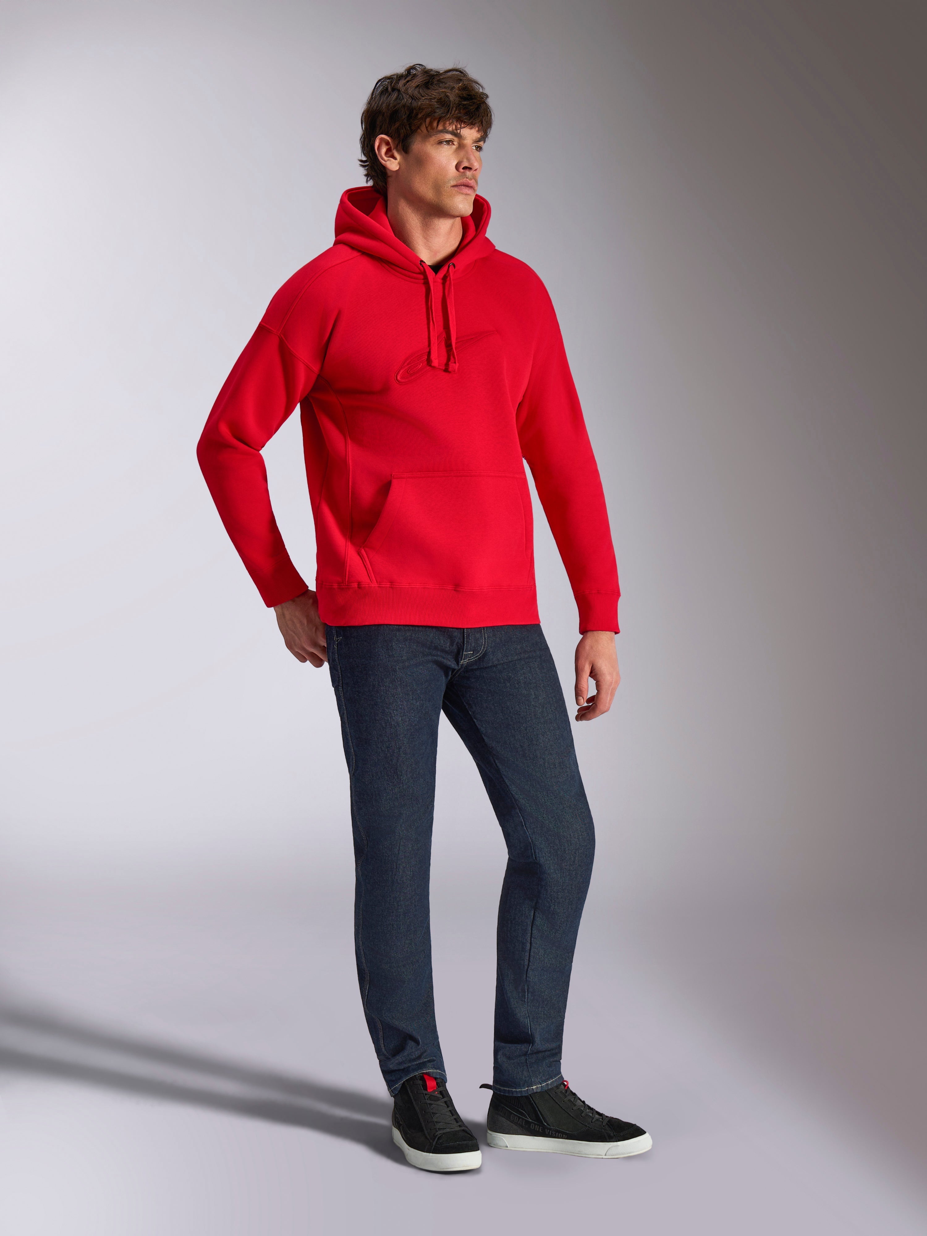 Alpinestars Emergent Oversized hættetrøje, rød, pullover fleece-hættetrøje med tonalt 3D-broderet logo, oversized pasform, båret af en model med mørkeblå jeans og sorte sneakers, afslappet motorsport-inspireret streetwear