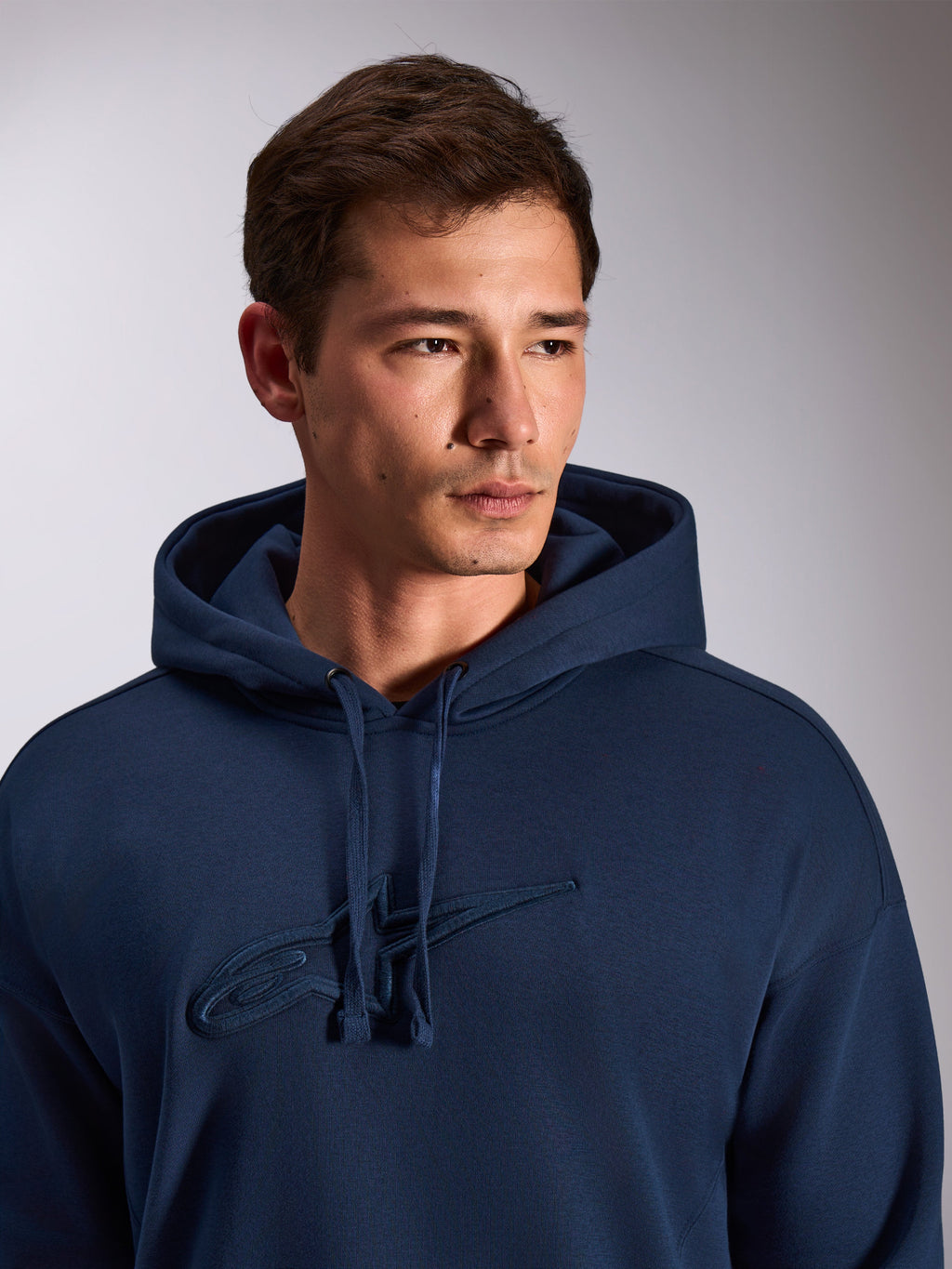 Alpinestars Emergent Oversized hættetrøje, navyblå, nærbillede af model iført pullover fleece-hættetrøje, med tonalt 3D-broderet Astars-logo på brystet, justerbar hætte med snøre, afslappet motorsport-streetwear
