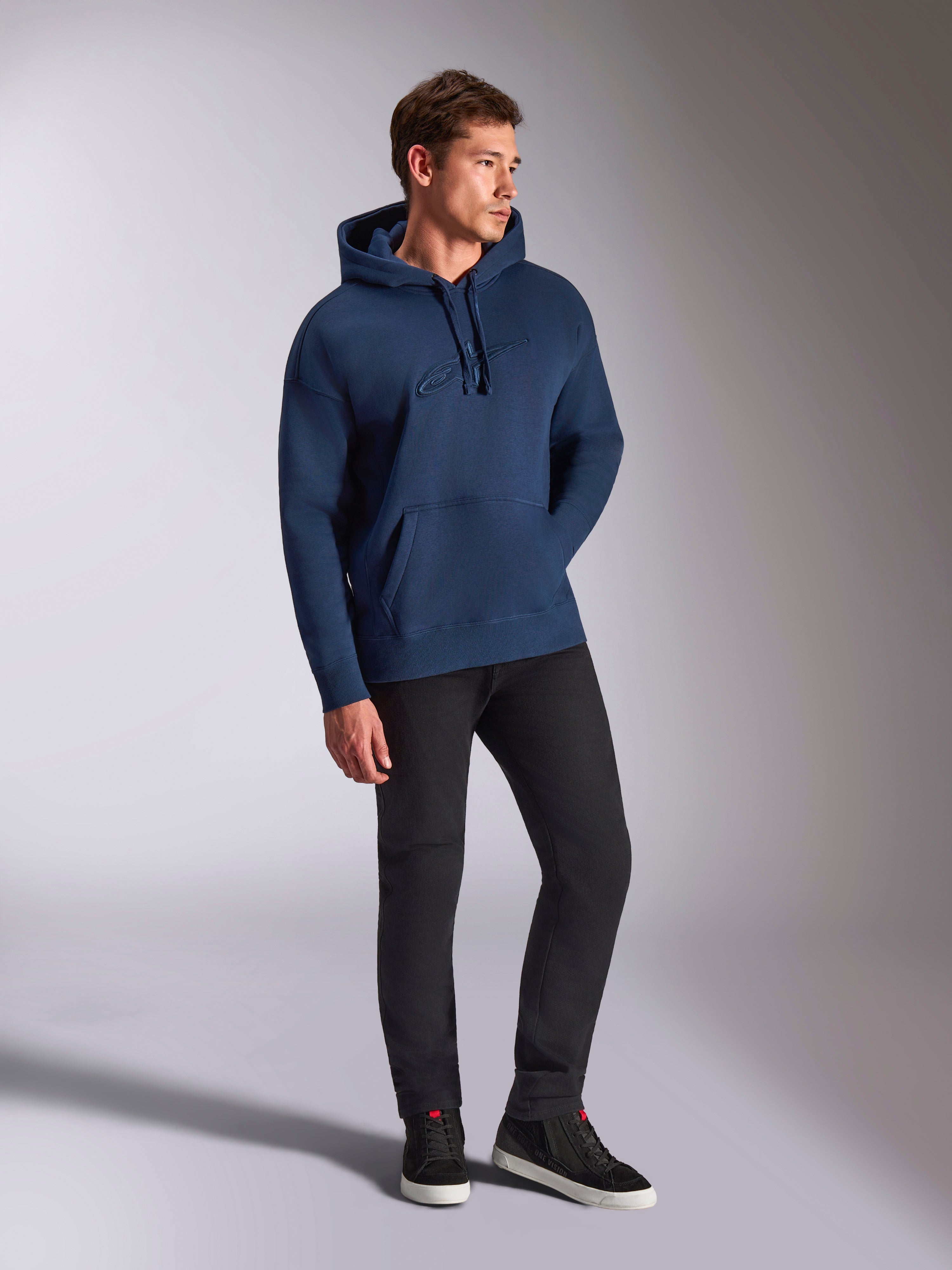 Alpinestars Emergent Oversized Hoodie, marineblå, bæres af en mandlig model med sorte bukser og sneakers, med 3D-broderet Astars-logo, kængurulomme og hætte med snøre, afslappet motorsport-inspireret streetwear.