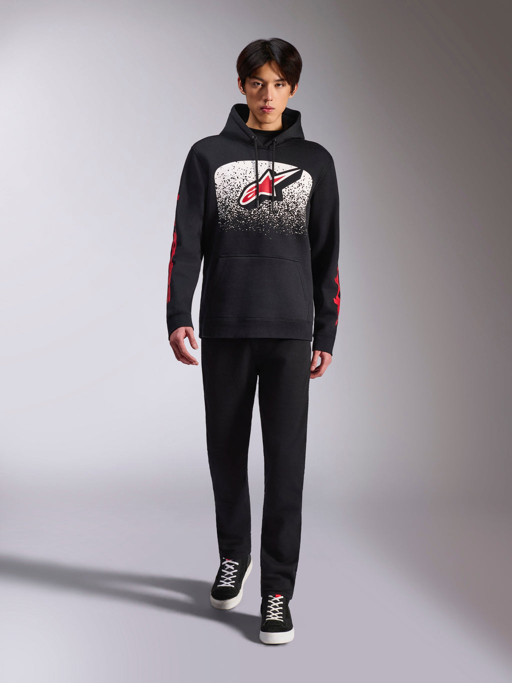 Alpinestars Obsessed hættetrøje, sort pullover-hættetrøje, stort hvidt og rødt Astars-logo med MX-graduering, røde word-logoer på ærmerne, båret af model i et helt sort afslappet outfit, motorsport-inspireret livsstilstøj