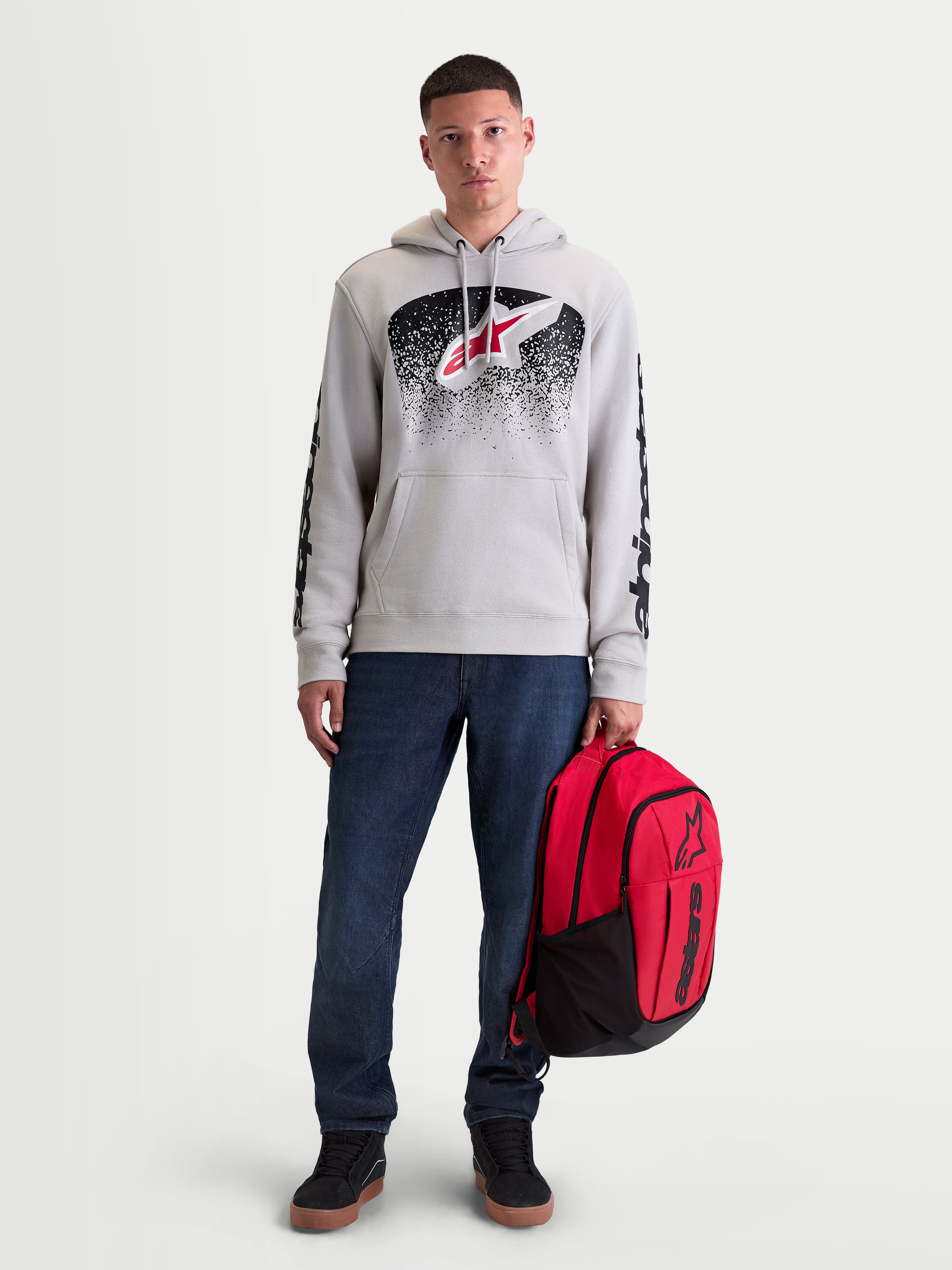 Alpinestars Obsessed Hættetrøje, herrehættetrøje i fleece til motorsport og fritid, cementgrå med rød og sort MX-inspireret gradientgrafik, branding på ærmerne, vist på mandlig model med rød rygsæk.