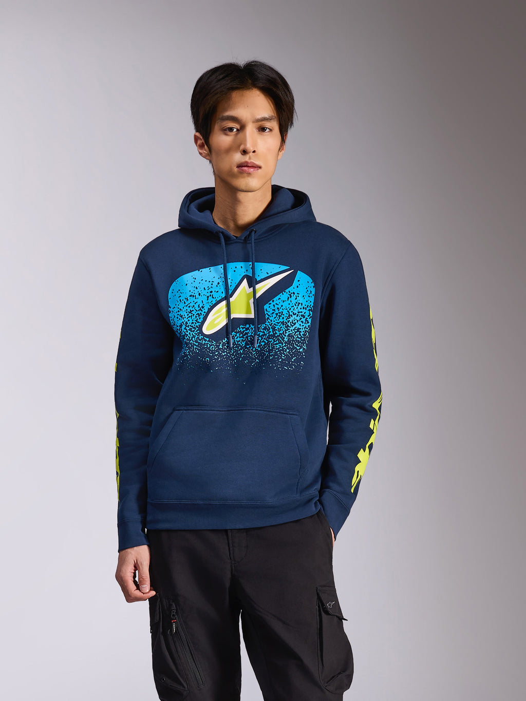 Alpinestars Obsessed-hættetrøje i Navy, båret af en mandlig model, med et limegrønt Astars-logo med lyseblå MX-graduering på brystet og gule wordmark-logoer på ærmerne, afslappet motorsport-inspireret streetwear