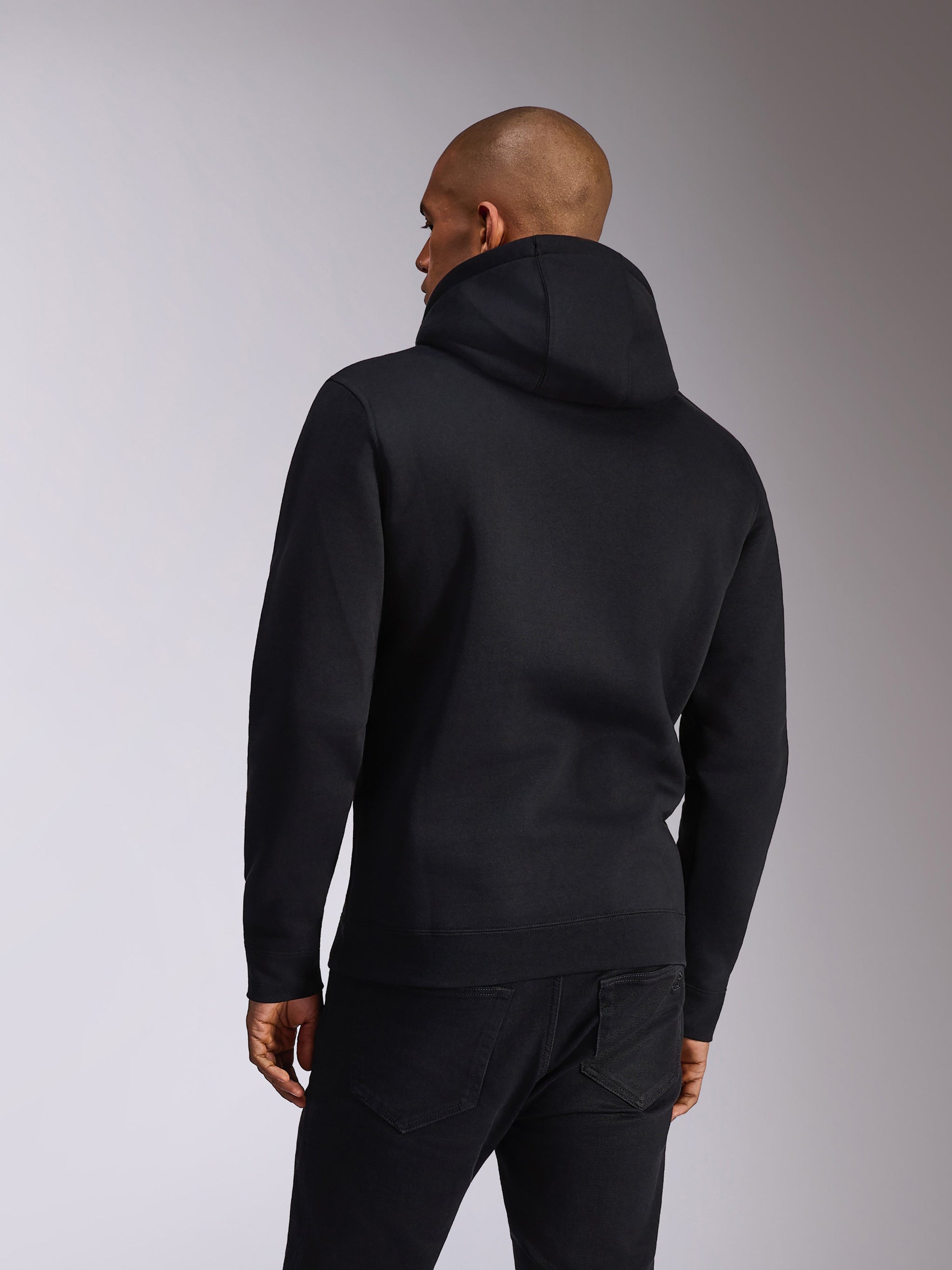 Alpinestars Ellipsoid Hoodie, Sort, set bagfra på en person iført en pullover-hættetrøje i fleece, lange ærmer med ribkanter og -søm, stylet med sorte jeans, afslappet motorsport-inspireret sportstøj