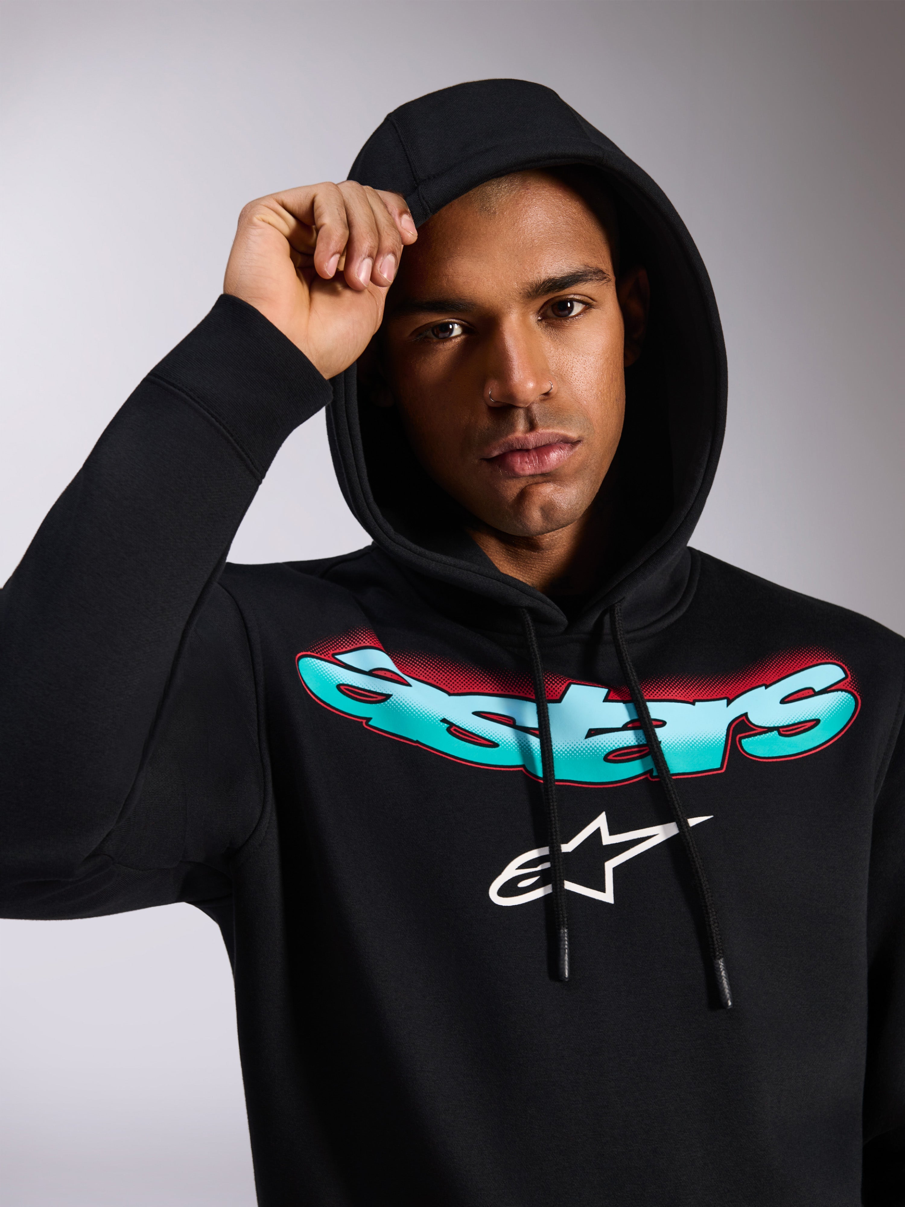 Alpinestars Ellipsoid Hoodie, black pullover fleece hoodie, featuring stylized teal and red color gradient Astars wordmark and white logo on chest, worn by model with hood up, casual motorsport-inspired streetwear