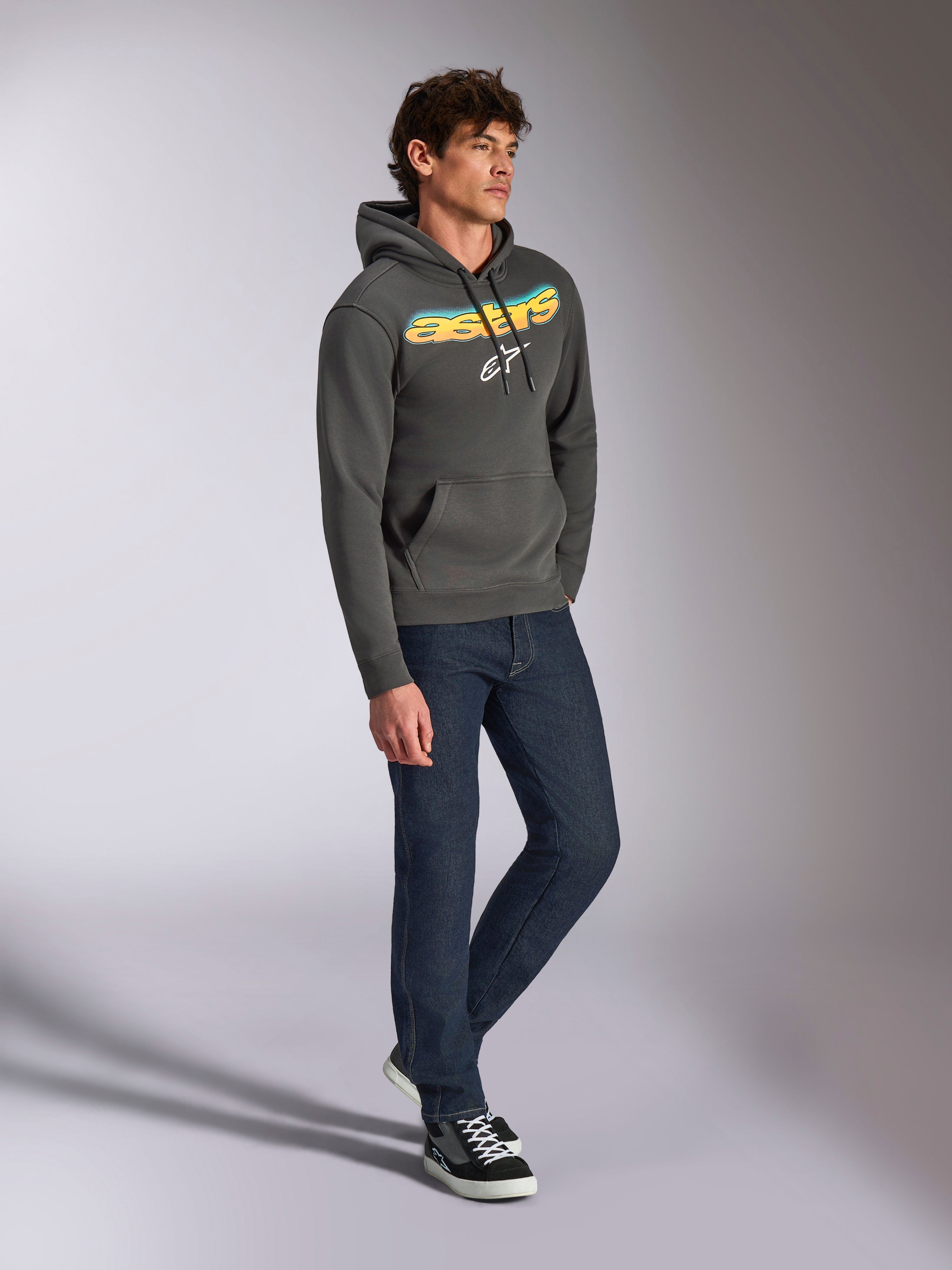 Alpinestars Ellipsoid hættetrøje, gunmetal grå pullover-fleece, buet Astars-tekst på fronten med blå og gul farveovergang og hvidt logo, stylet med mørke jeans og sorte sneakers, afslappet streetwear.