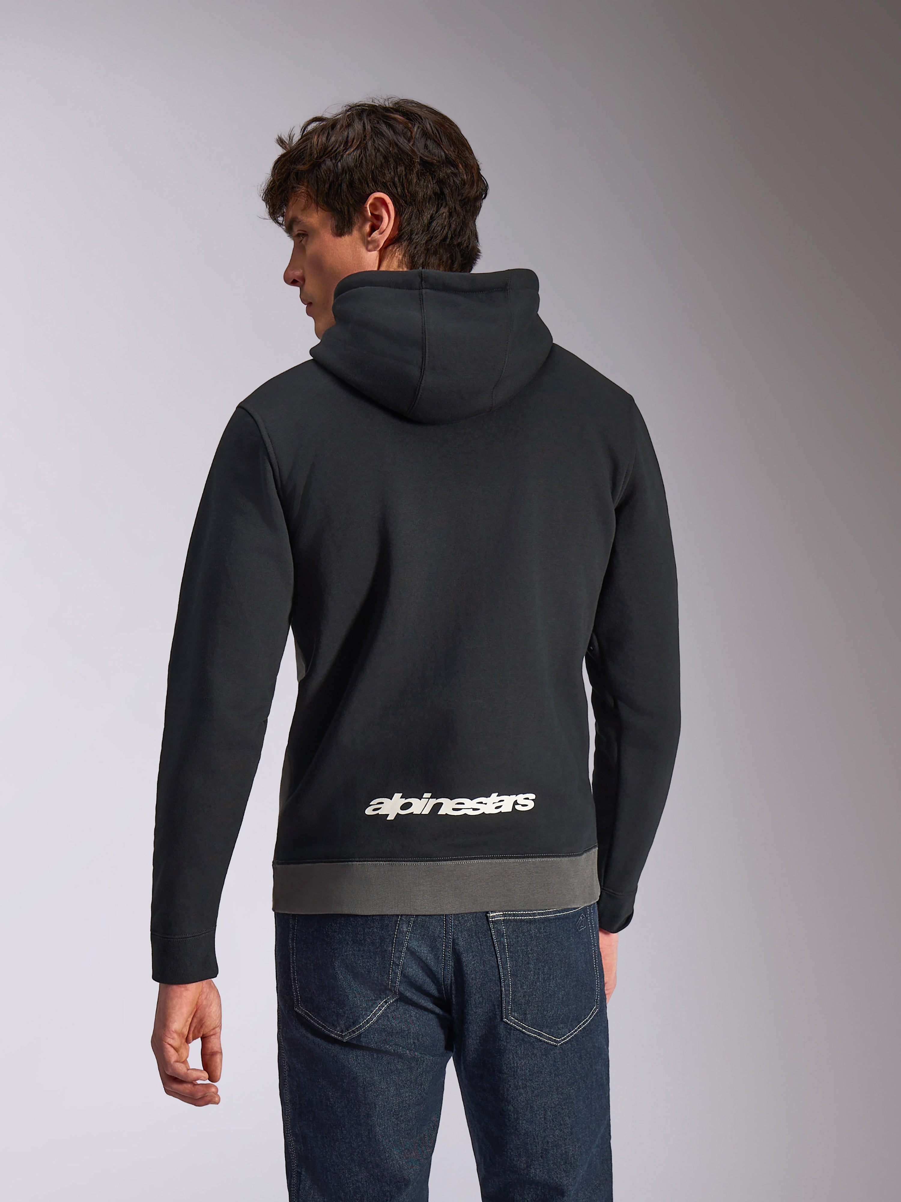 Alpinestars Parallel hættetrøje, casual sportswear hættetrøje, sort og gunmetal, set bagfra med hvidt Alpinestars-logo nederst på ryggen, farveblokket design med grå kant, båret med mørke denimjeans