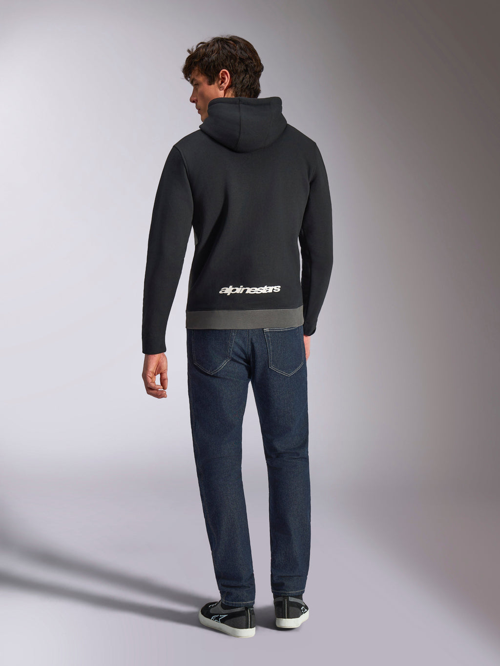 Alpinestars Parallel hættetrøje, mand set bagfra iført sort og gunmetal farveblokket pullover-fleece, hvidt Alpinestars ordlogo på den nederste del af ryggen, mørkeblå jeans, afslappet motorsport-inspireret streetwear