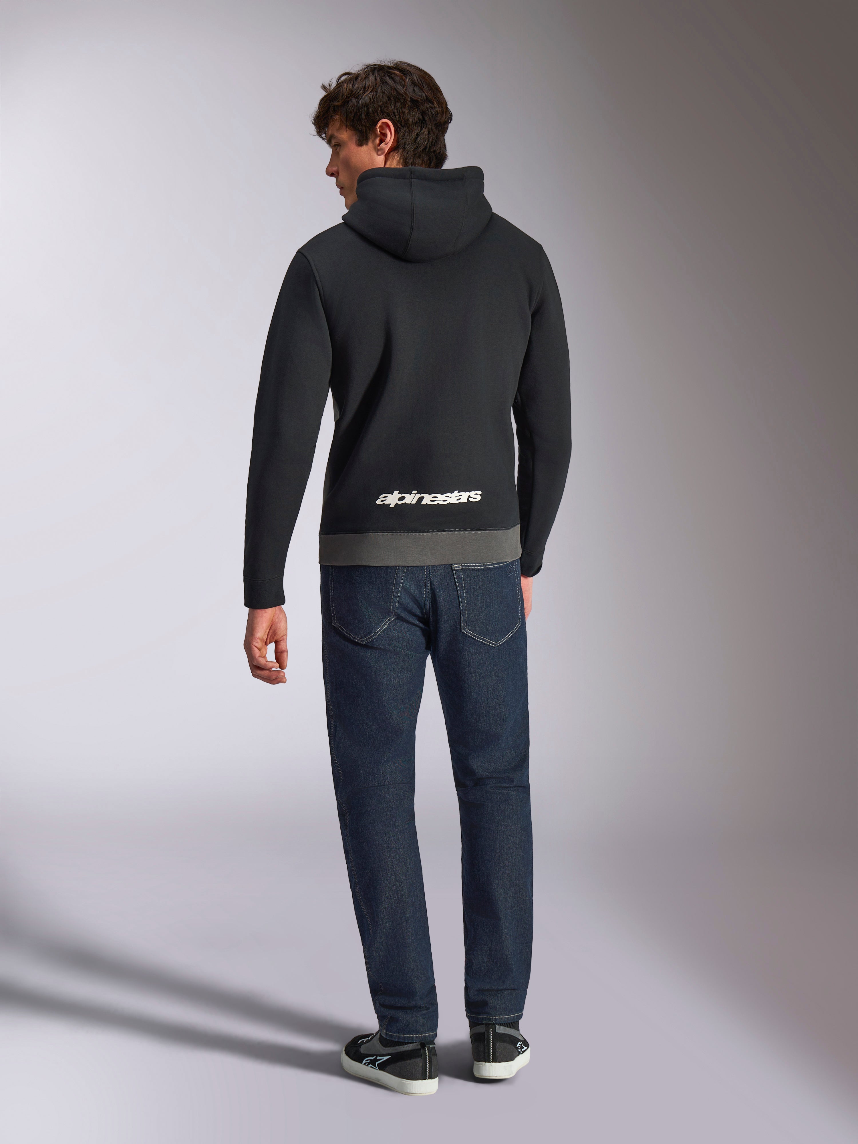 Alpinestars Parallel hættetrøje, mand set bagfra iført sort og gunmetal farveblokket pullover-fleece, hvidt Alpinestars ordlogo på den nederste del af ryggen, mørkeblå jeans, afslappet motorsport-inspireret streetwear