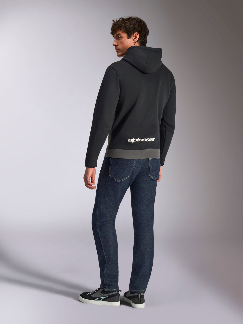 Alpinestars Parallel Hoodie, set bagfra på model der bærer den sorte og gunmetal-grå pullover, med hvidt Alpinestars-logo nederst på ryggen, stylet med mørkeblå jeans, afslappet motorsport-inspireret streetwear