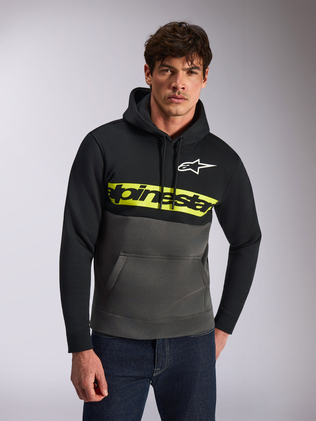 Alpinestars Parallel hættetrøje, afslappet streetwear, farveblokket sort og koksgrå, klar gul bryststribe med Alpinestars-ordlogo, hvidt Astars-logo, båret af en model med blå jeans, moderne motorsport-inspireret design
