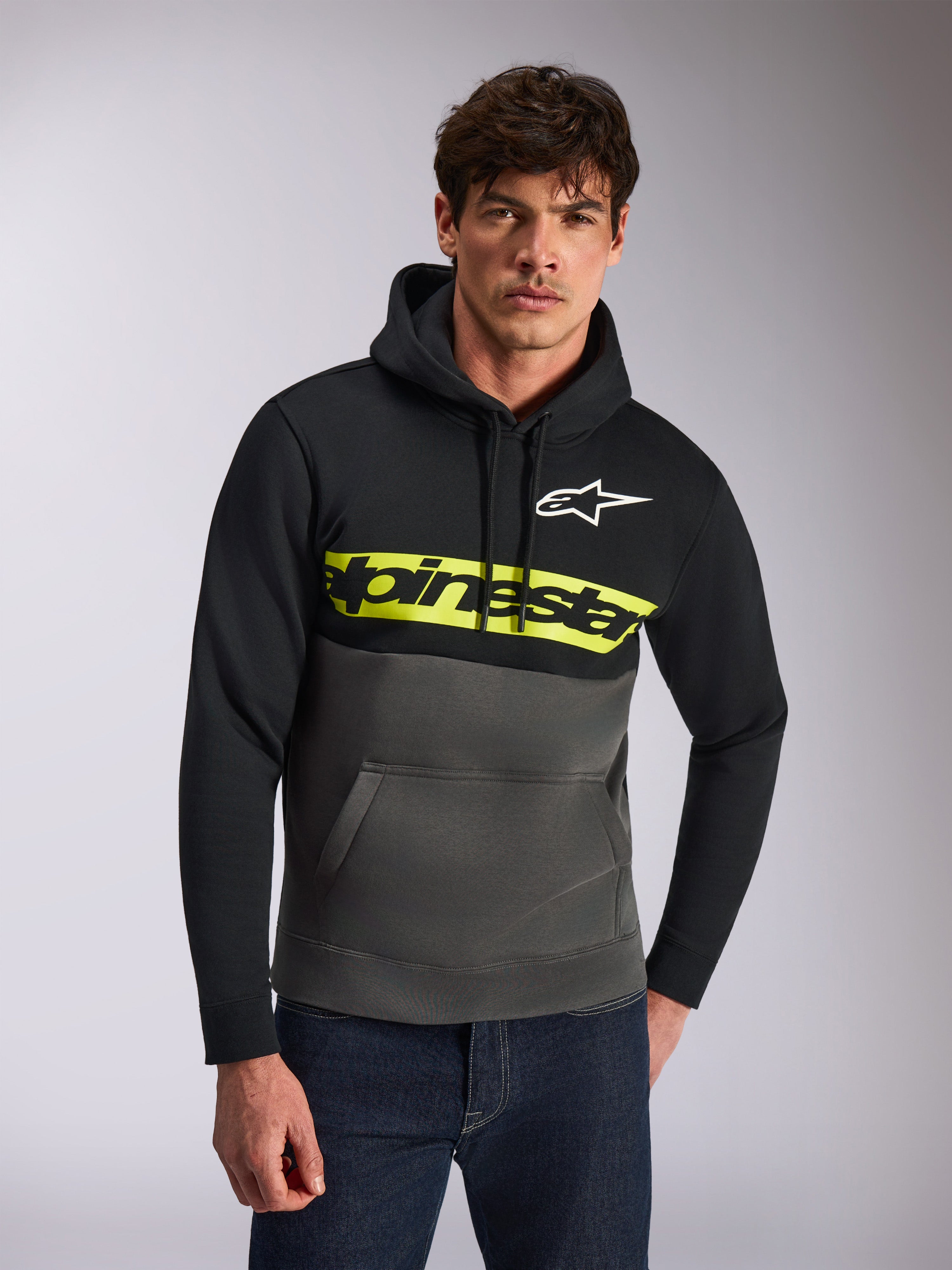 Alpinestars Parallel hættetrøje, afslappet streetwear, farveblokket sort og koksgrå, klar gul bryststribe med Alpinestars-ordlogo, hvidt Astars-logo, båret af en model med blå jeans, moderne motorsport-inspireret design