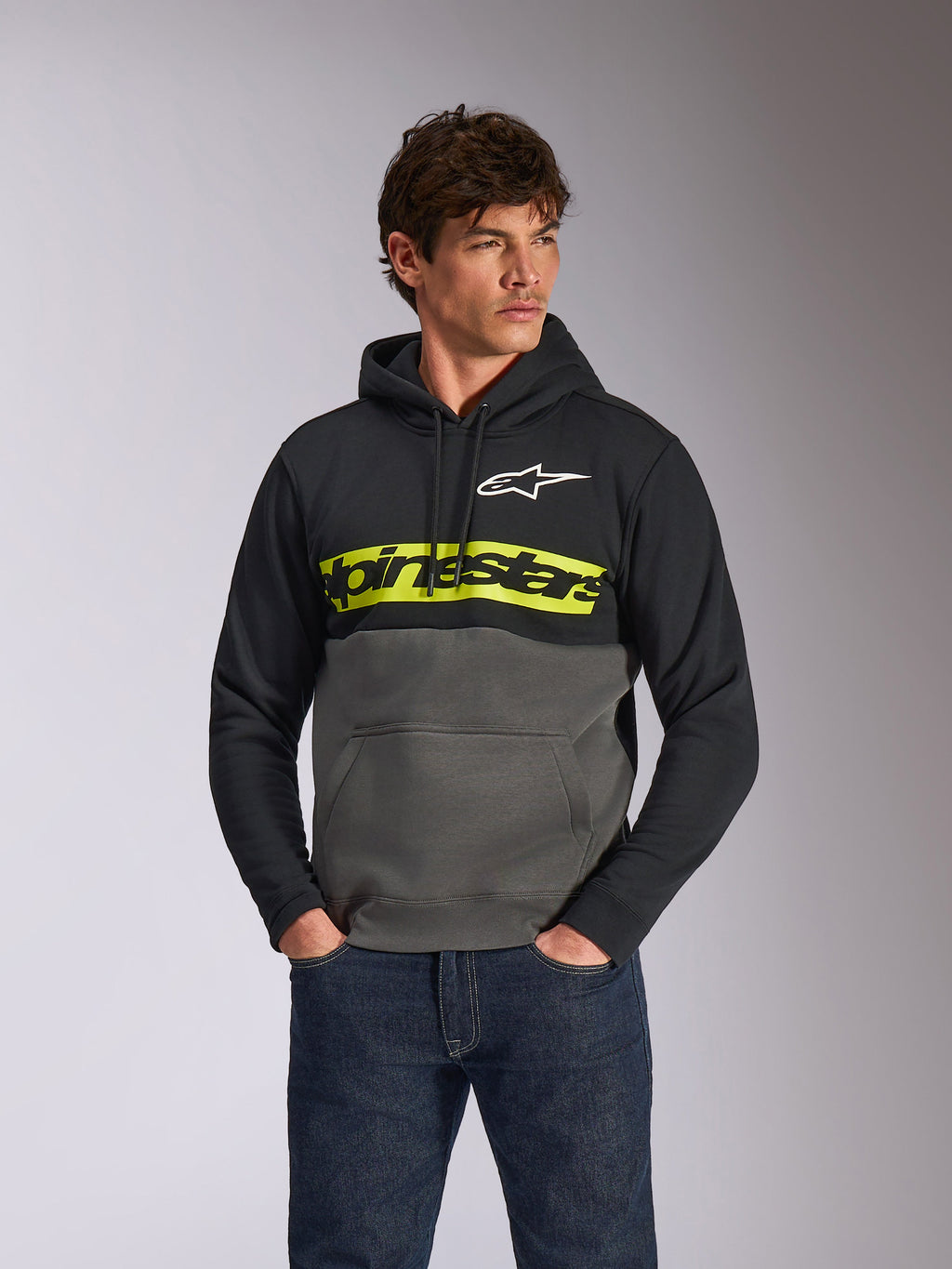 Alpinestars Parallel hættetrøje, afslappet farveblokket fleece-pullover, sort og gunmetal med en markant gul bryststribe, Alpinestars-tekstlogo og hvidt stjernelogo, båret af en mandlig model med mørkeblå jeans