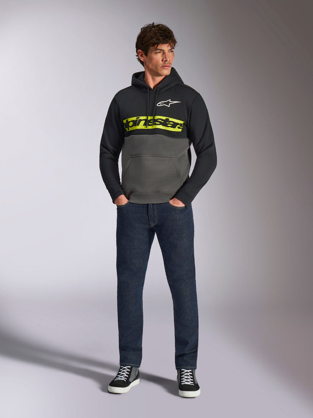Alpinestars Parallel-hættetrøje, farveblokket pullover, sort og gunmetal-grå, skarpt gult Alpinestars-tekstlogo og hvidt stjernelogo på brystet, båret af en mandlig model med mørkeblå jeans og sorte sneakers, afslappet motorsport-streetwear