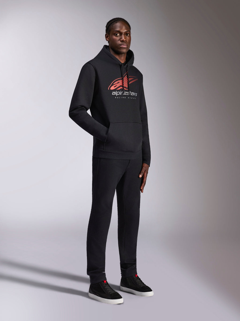 Alpinestars Pulse Hoodie, sort pullover hættetrøje, med rødt og hvidt Astars-logo med speed line-fade og Racing Ahead-tekst, båret med matchende sorte bukser og sneakers, afslappet motorsport-inspireret streetwear.