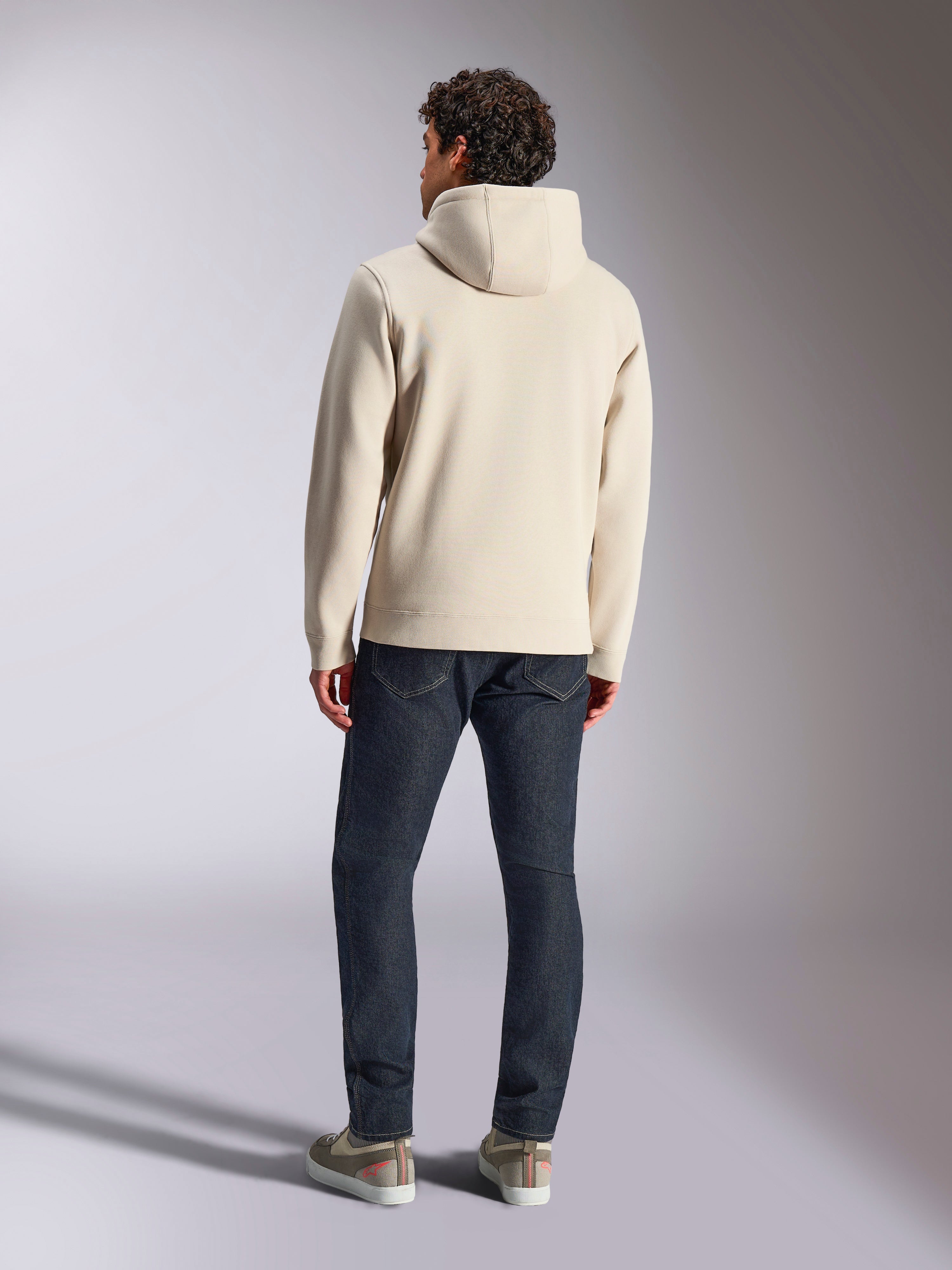 Alpinestars Pulse Hoodie, Off White, set bagfra af en mand iført en afslappet pullover fleece-hættetrøje med mørke jeans og sneakers, moderne sportswear-design