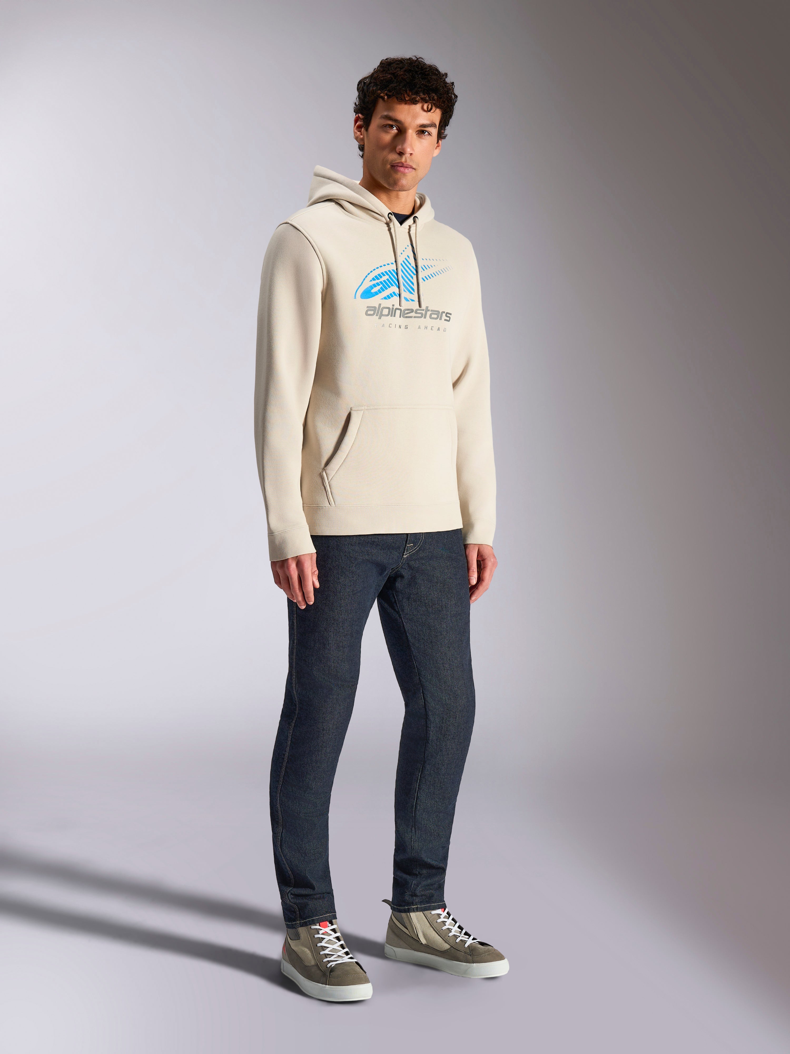 Alpinestars Pulse Hoodie, Off White afslappet streetwear, fleece-hættetrøje med blåt og gråt Astars-logo med speed line-fade, kængurulomme, båret af model med mørke denimjeans og high-top sneakers.