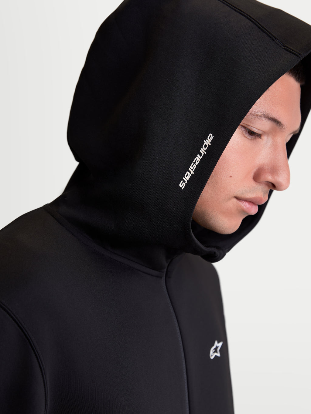 Alpinestars Cypher hættetrøje, herre sportstøj lynlåshættetrøje, sort, nærbillede på model der viser den høje hætte med hvidt Alpinestars-ordlogo og teknisk tri-blend fleece.