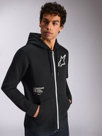Alpinestars Transverse hættetrøje, afslappet sportstøj, sort med hvid lynlåsdetalje, hætte med snoretræk, Alpinestars stjerne-logo på brystet, 'One Goal One Vision'-grafik, båret af mandlig model med mørkt krøllet hår