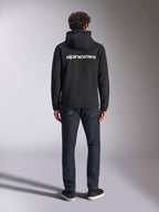 Alpinestars Transverse hættetrøje, casual motorsport streetwear, sort med hvidt Alpinestars-tekstlogo på ryggen, lange ærmer, ribkant forneden og ved manchetterne, set bagfra med mørke denimjeans
