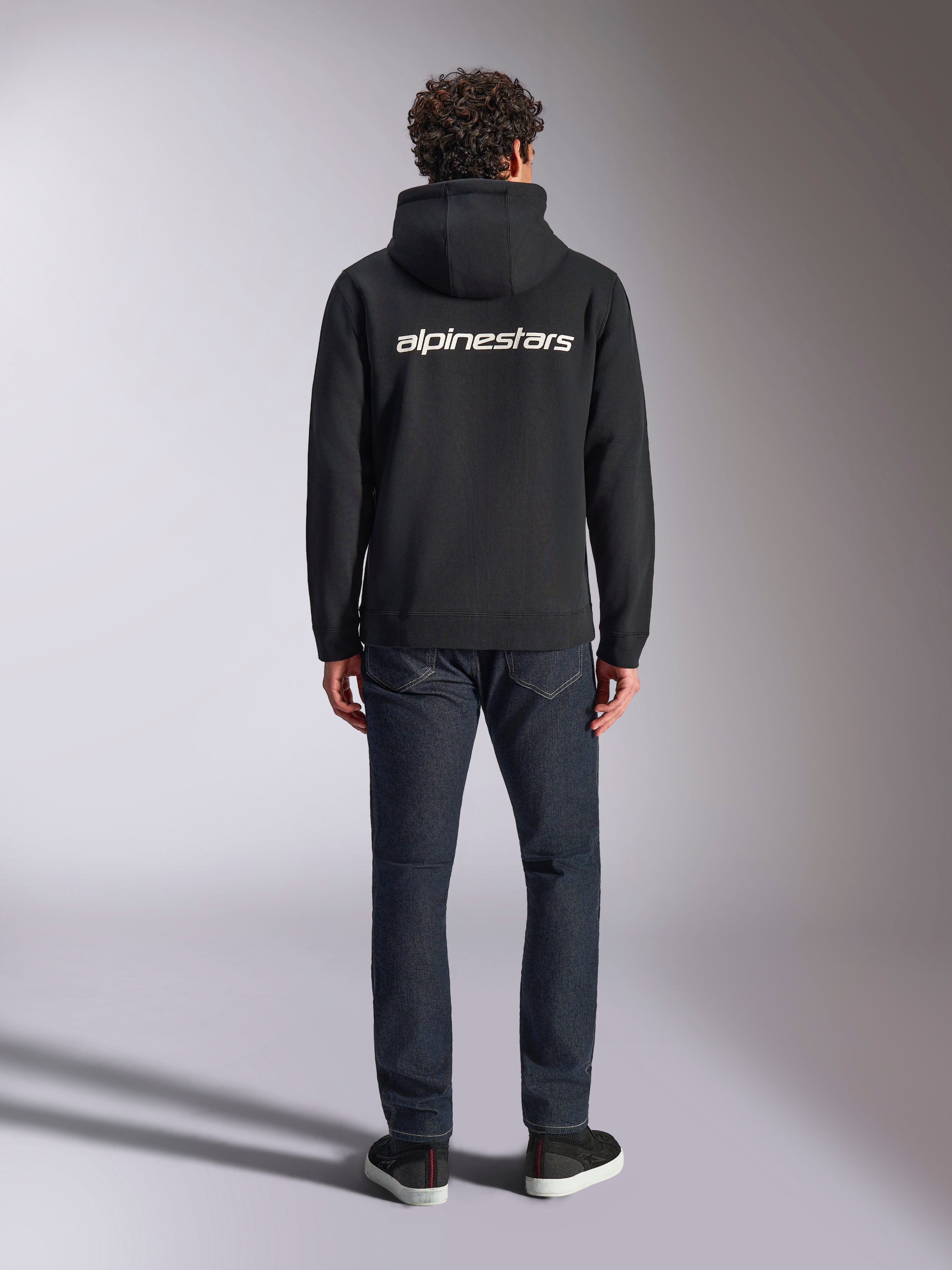 Alpinestars Transverse hættetrøje, casual motorsport streetwear, sort med hvidt Alpinestars-tekstlogo på ryggen, lange ærmer, ribkant forneden og ved manchetterne, set bagfra med mørke denimjeans