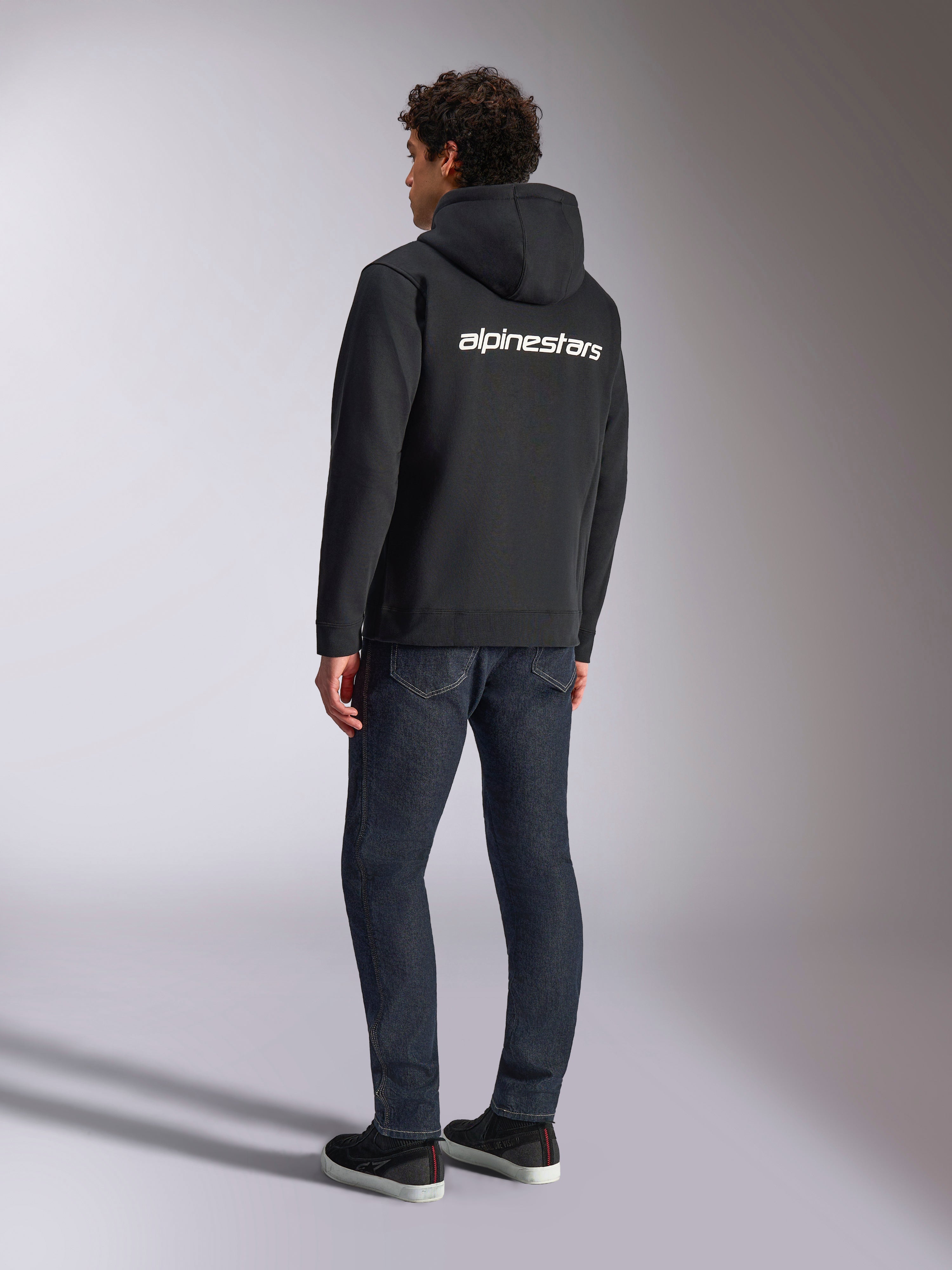 Alpinestars Transverse Hoodie, sort fleecehættetrøje med lynlås, set bagfra med hvidt Alpinestars-tekstlogo over skuldrene, båret med mørkeblå jeans og sorte sneakers, afslappet motorsport-inspireret streetwear