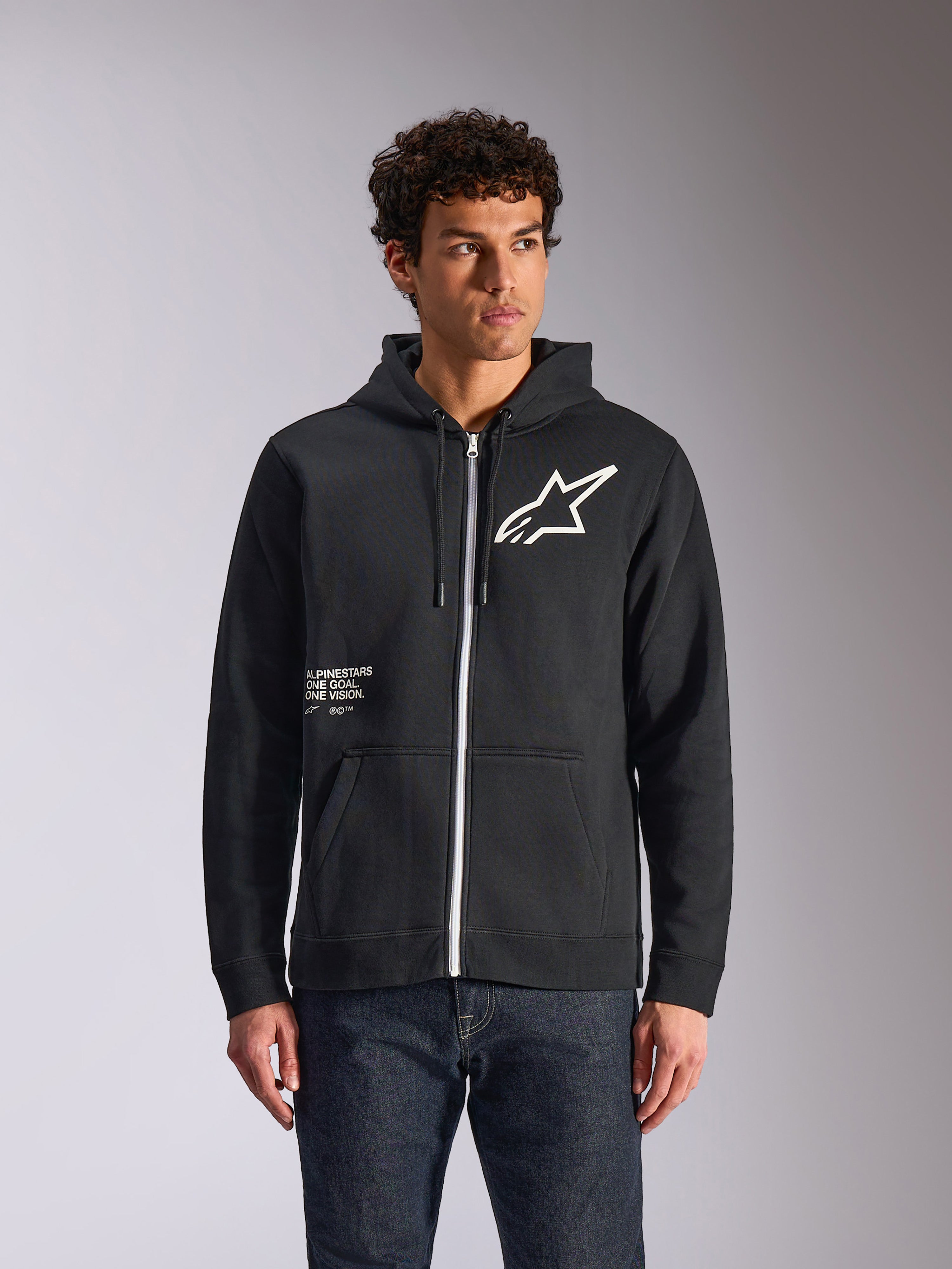 Alpinestars Transverse Hoodie, sort fleece-hættetrøje med lynlås foran, med hvidt Astars-logo og One Goal-grafik på forsiden, båret af en model med mørke jeans, afslappet motorsport-inspireret streetwear