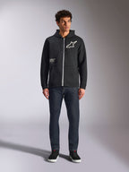 Alpinestars Transverse Hoodie, afslappet fleece med lynlås foran, sort, hvidt Astars-logo på brystet og 'One Goal'-print på forsiden, hætte med snøre, kombineret med mørke jeans og sorte sneakers, lifestyle motorsport streetwear