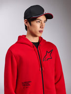 Alpinestars Transverse-hættetrøje, rød fleece med lynlås foran, sort Astars-logo på brystet, 'One Goal. One Vision.'-tekst nederst på forsiden, afslappet motorsport-streetwear, båret af en model med en sort kasket