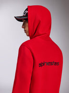 Alpinestars Transverse Hoodie, rød fleece-hættetrøje, set fra siden bagfra, der viser det sorte Alpinestars-navnetræk på ryggen, båret med hætten oppe og en sort kasket, afslappet motorsport-inspireret streetwear