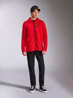 Alpinestars Transverse hættetrøje, rød fleece-hættetrøje med gennemgående lynlås, sort Astars-logo på brystet og One Goal-print på siden, båret af mandlig model med sorte bukser og kasket, afslappet motorsport-inspireret streetwear