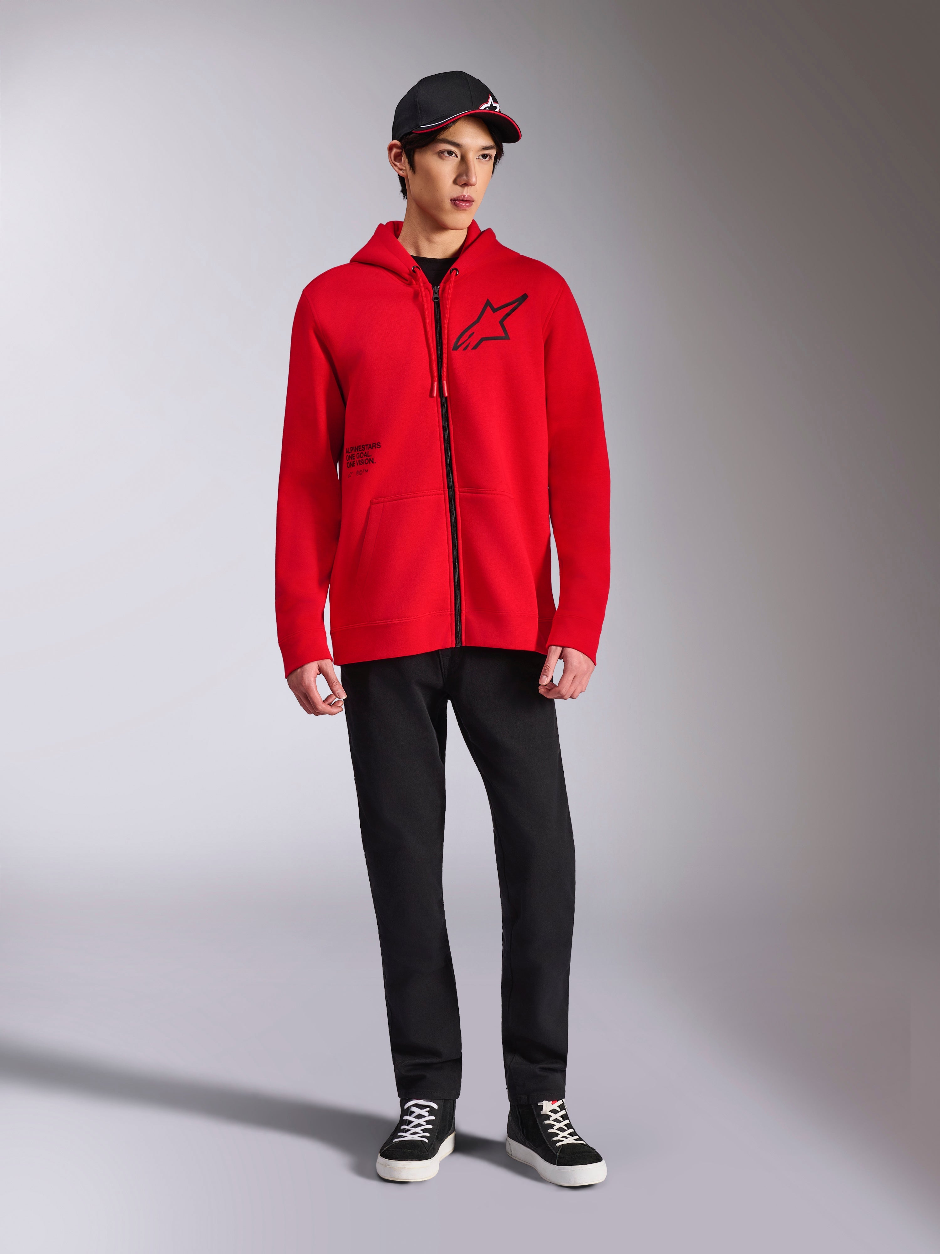 Alpinestars Transverse hættetrøje, rød fleece-hættetrøje med gennemgående lynlås, sort Astars-logo på brystet og One Goal-print på siden, båret af mandlig model med sorte bukser og kasket, afslappet motorsport-inspireret streetwear