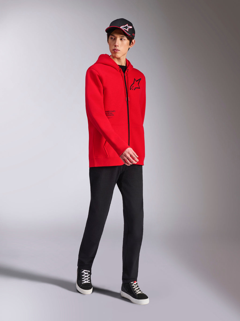 Alpinestars Transverse hættetrøje i rød, mandlig model iført fleecehættetrøje med lynlås, sort Astars-logo og tekstprint, stylet med sorte bukser og kasket, afslappet motorsport-streetwear