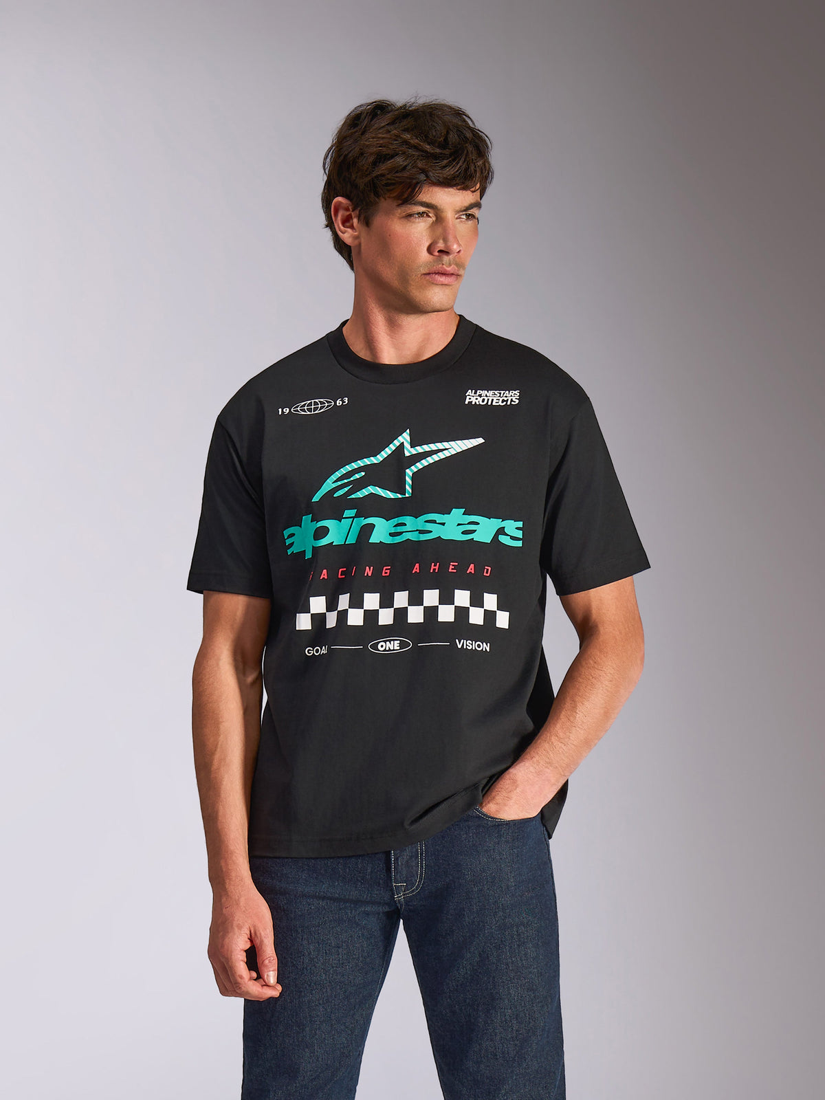 Alpinestars Aplomb Oversized CSF Tee, kortærmet T-shirt, sort, med et stort teal og hvidt Astars-kombinationslogo med ternet grafik, båret af mandlig model med mørkeblå jeans, afslappet motorsport-streetwear