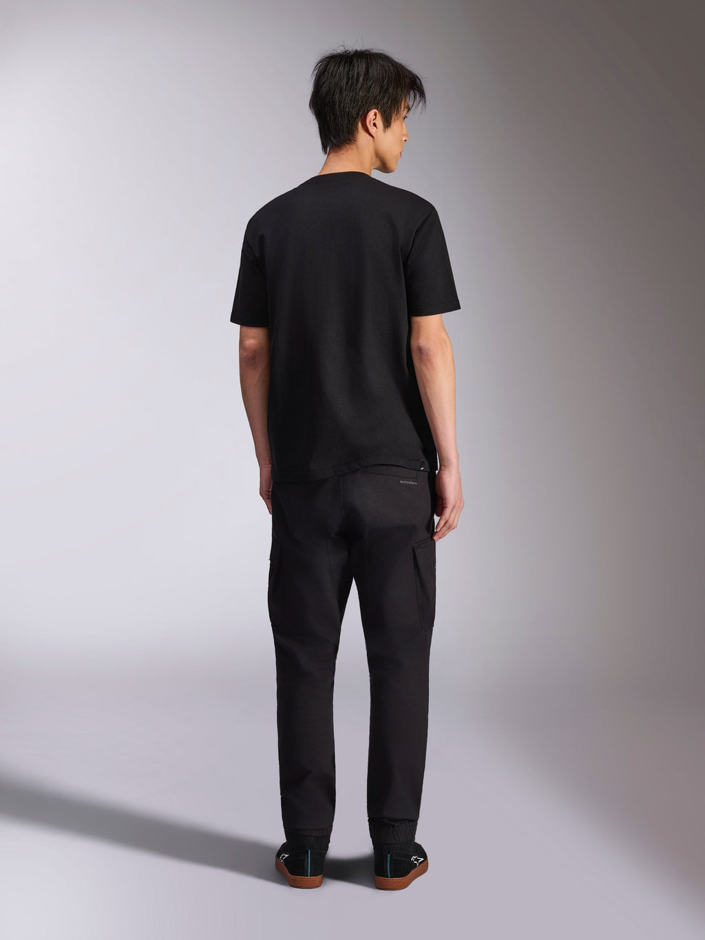 Alpinestars Noka CSF Tee, kortærmet t-shirt, sort, model set bagfra iført t-shirt med sorte bukser og sneakers, afslappet motorsport-streetwear-stil